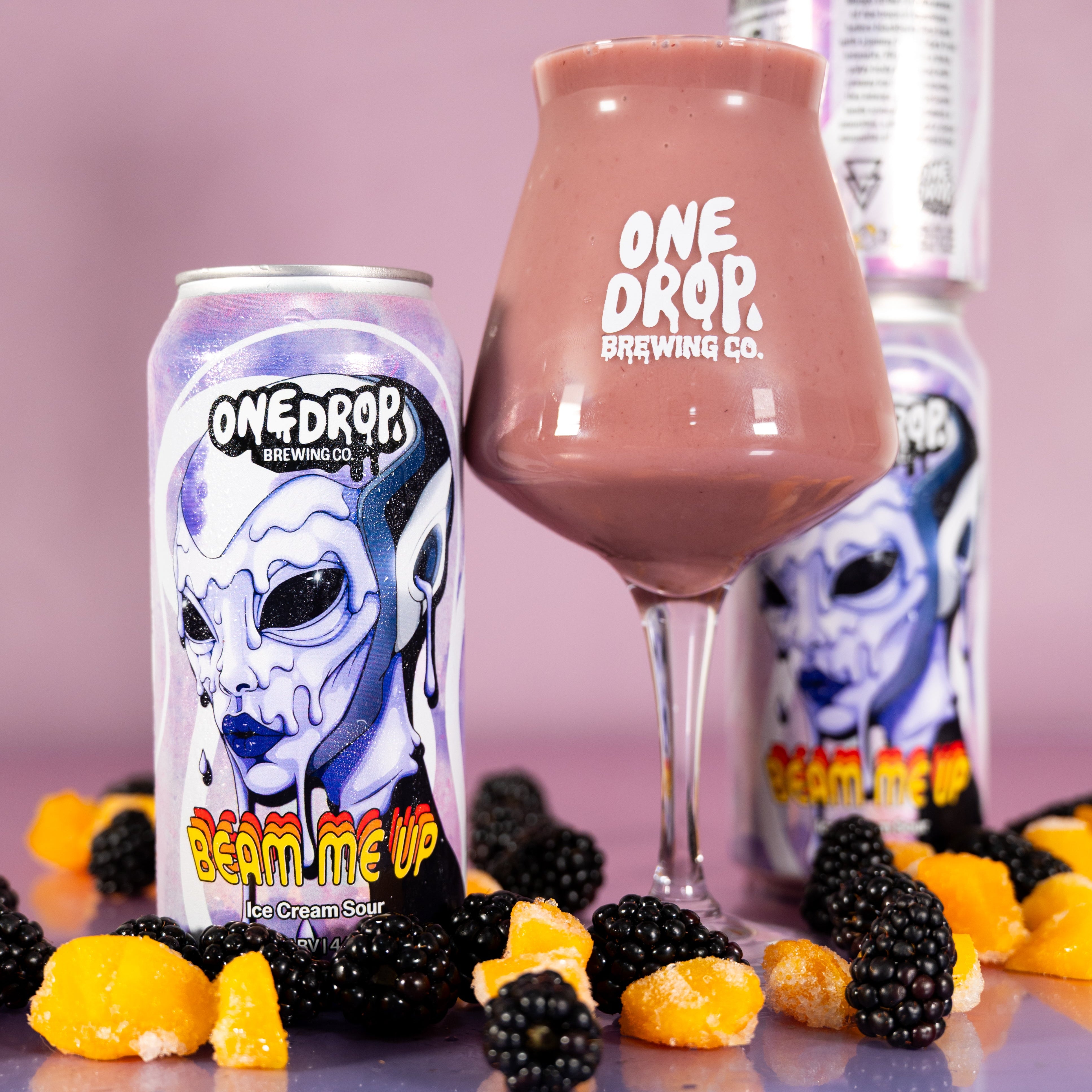 A beverage product setup on a reflective blue surface against a pink-lavender studio background. A can labeled ONE DROP BREWING CO.