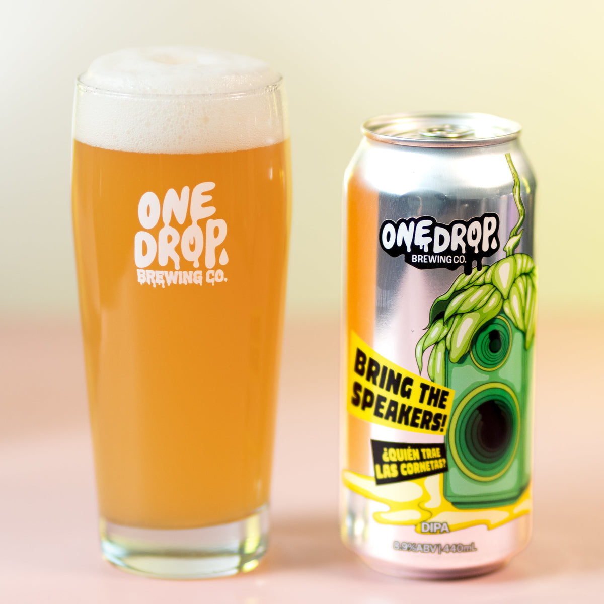 A glass of golden beer with white foam and a can of One Drop Brewing Co. 'Bring the Speaker!' IPA next to it on a pink surface against a pale yellow background.