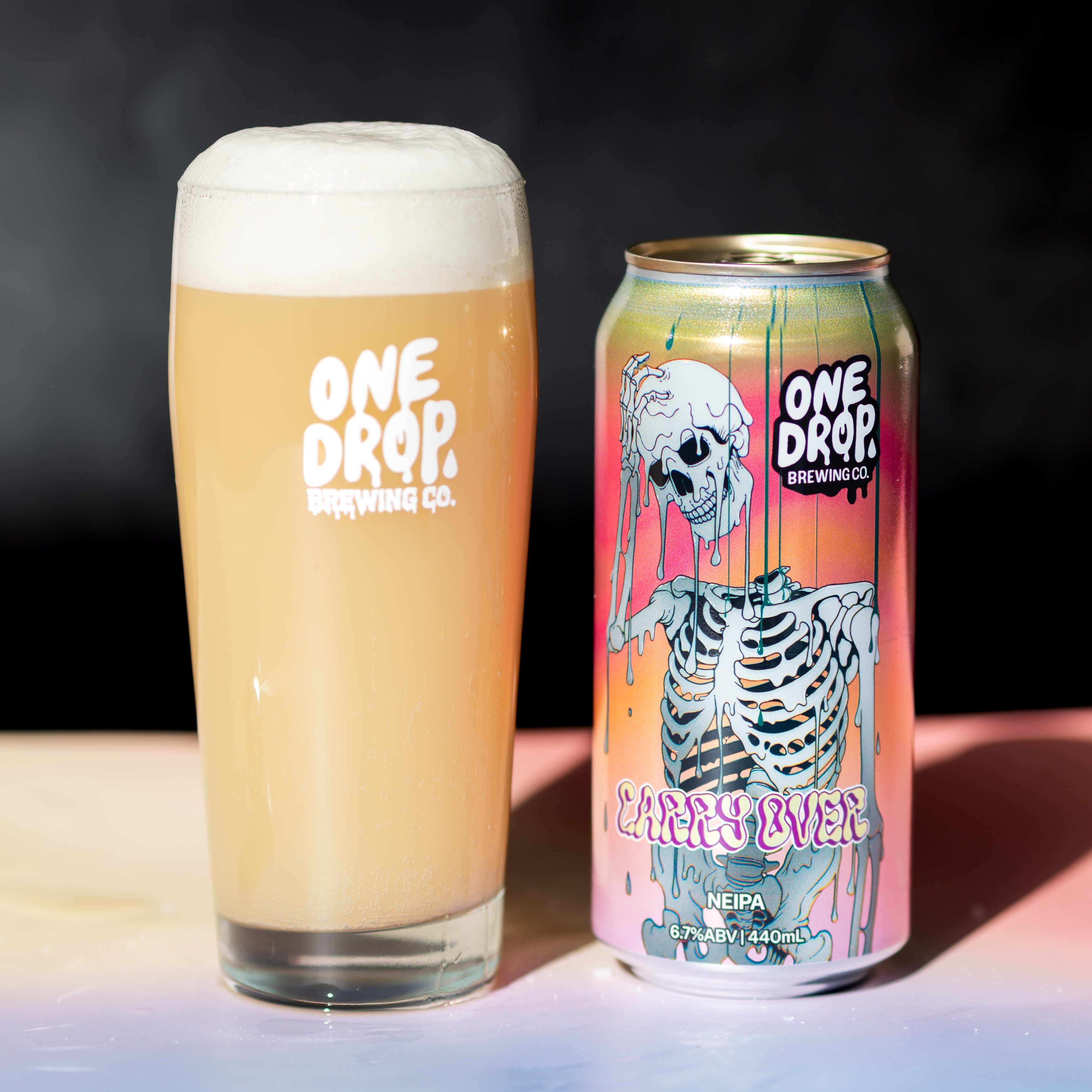 A frothy pint of One Drop Brewing Co. beer in a clear glass next to a matching illustrated can, both branded One Drop, set on a surface against a dark background.