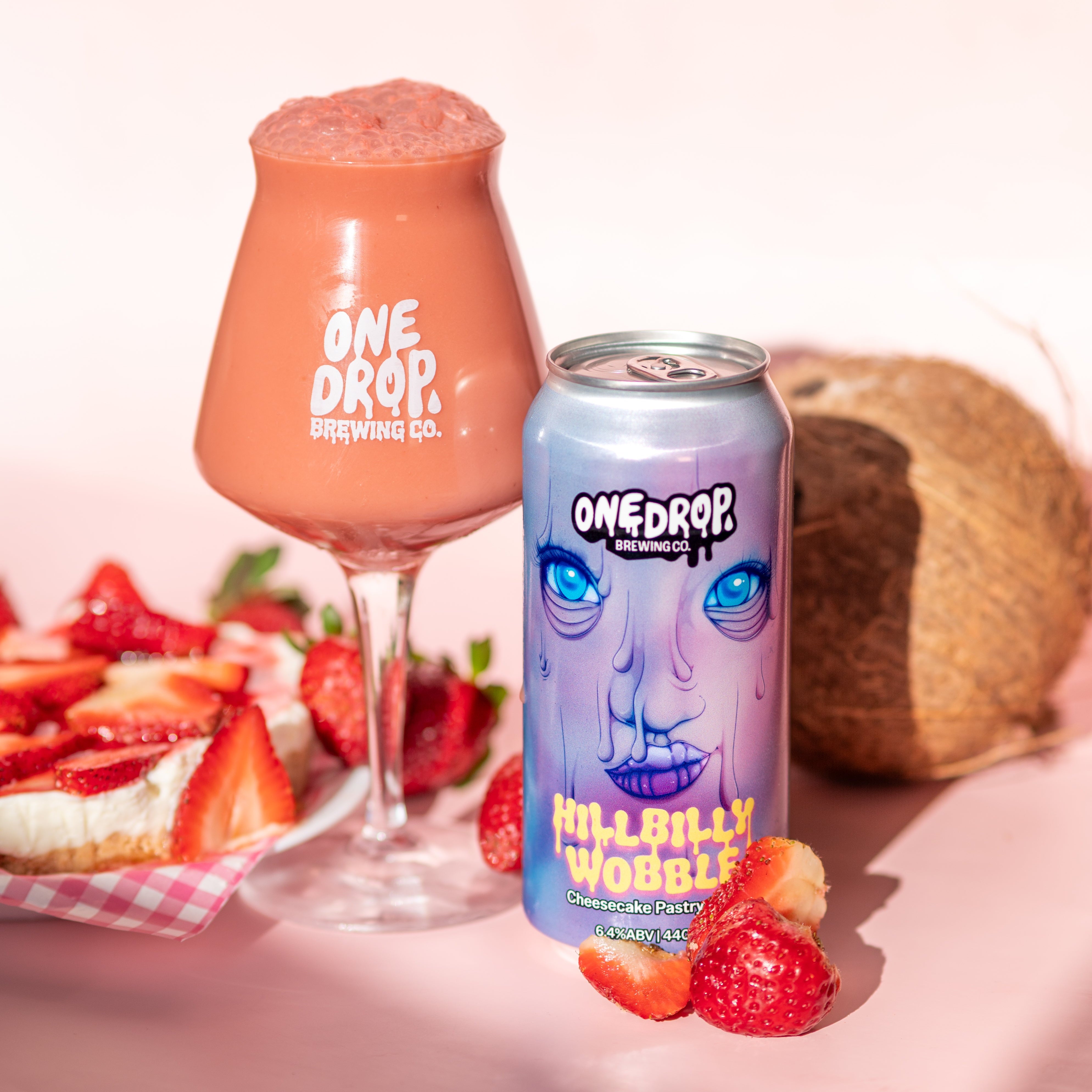 A can of One Drop Brewing Kill Billy Wobbie beer and a glass of the pink beverage, styled with strawberries and a coconut on a pink background.