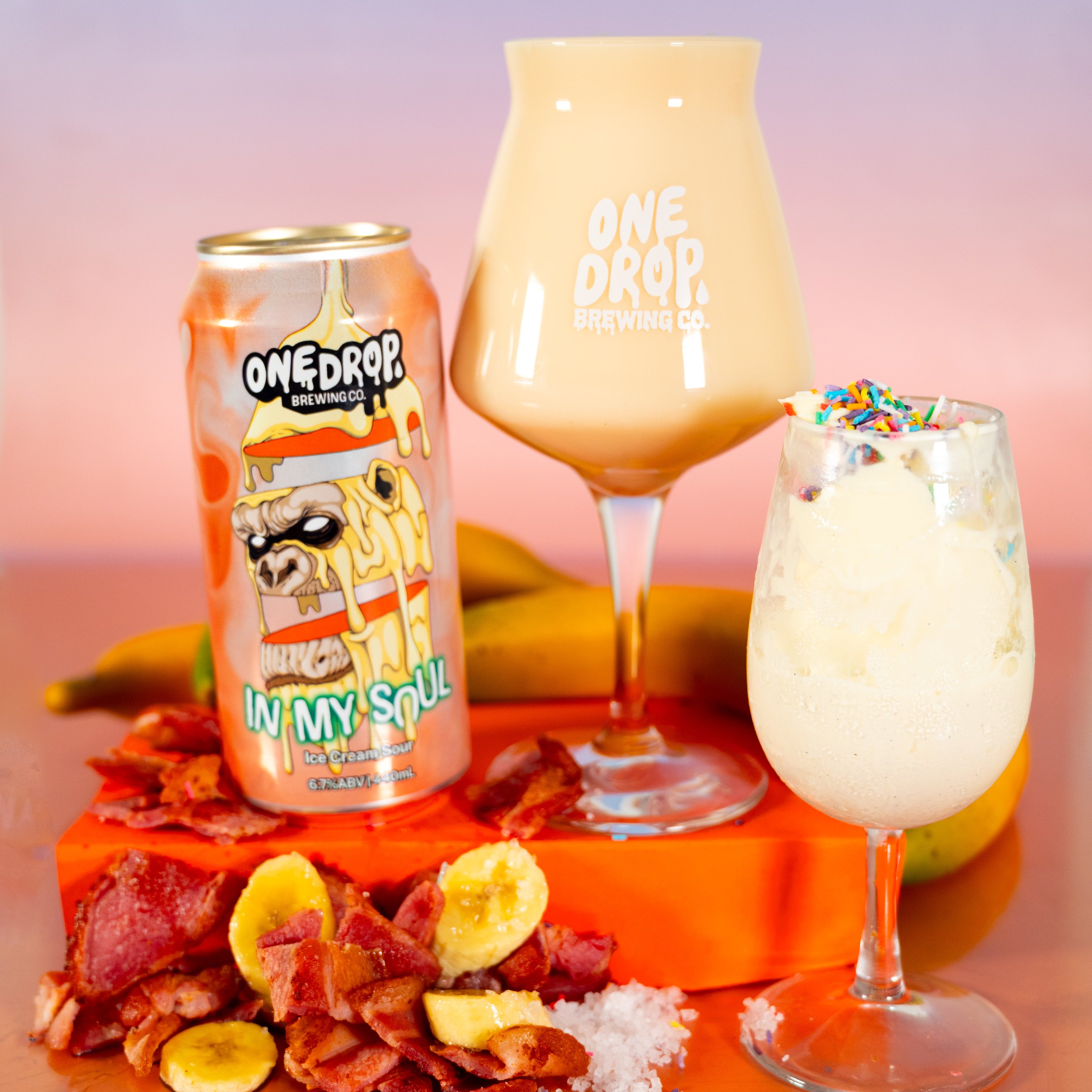 A beverage product display featuring a can of One Drop Brewing Co. 'In My Soul' banana malt beverage alongside two glasses of the drink, one in a tulip glass and one in a coupe glass with sprinkles, set on an orange platform with banana slices and bacon