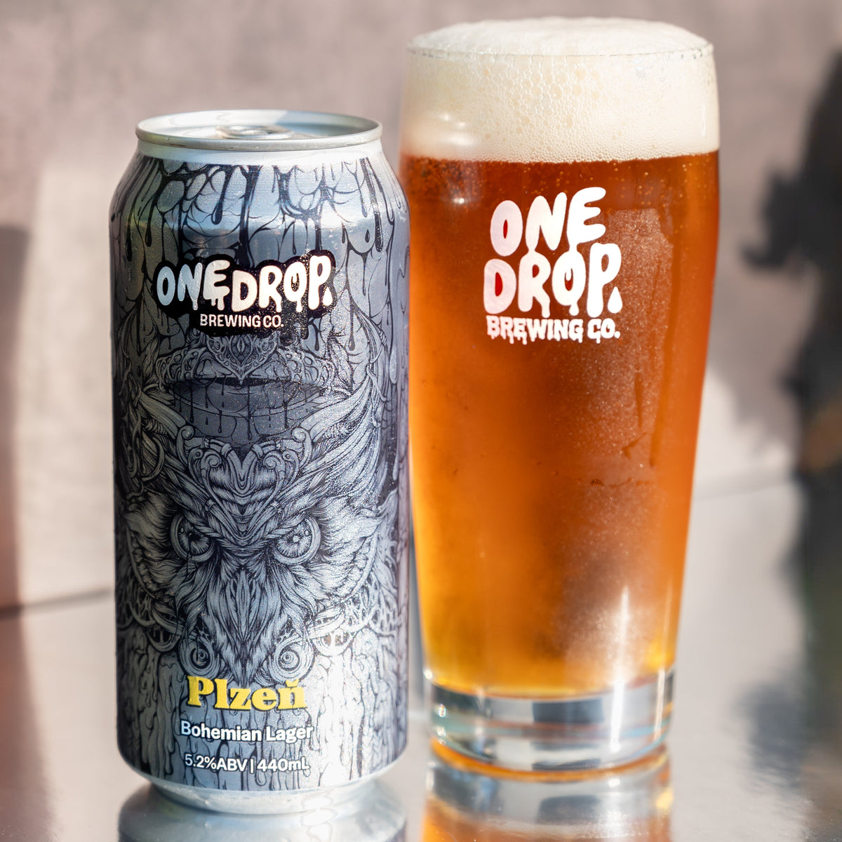 A can and a glass of One Drop Brewing Co. Plzen Bohemian Lager beer on a reflective metallic surface against a textured gray background.