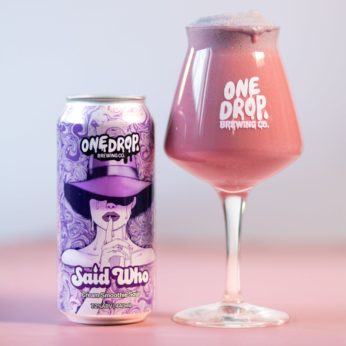 A can and a stemmed glass of One Drop Brewing Co. Said Who beer, showing the purple-labeled can and a pinkish-purple beverage in a tulip glass on a pink surface against a light gray background.