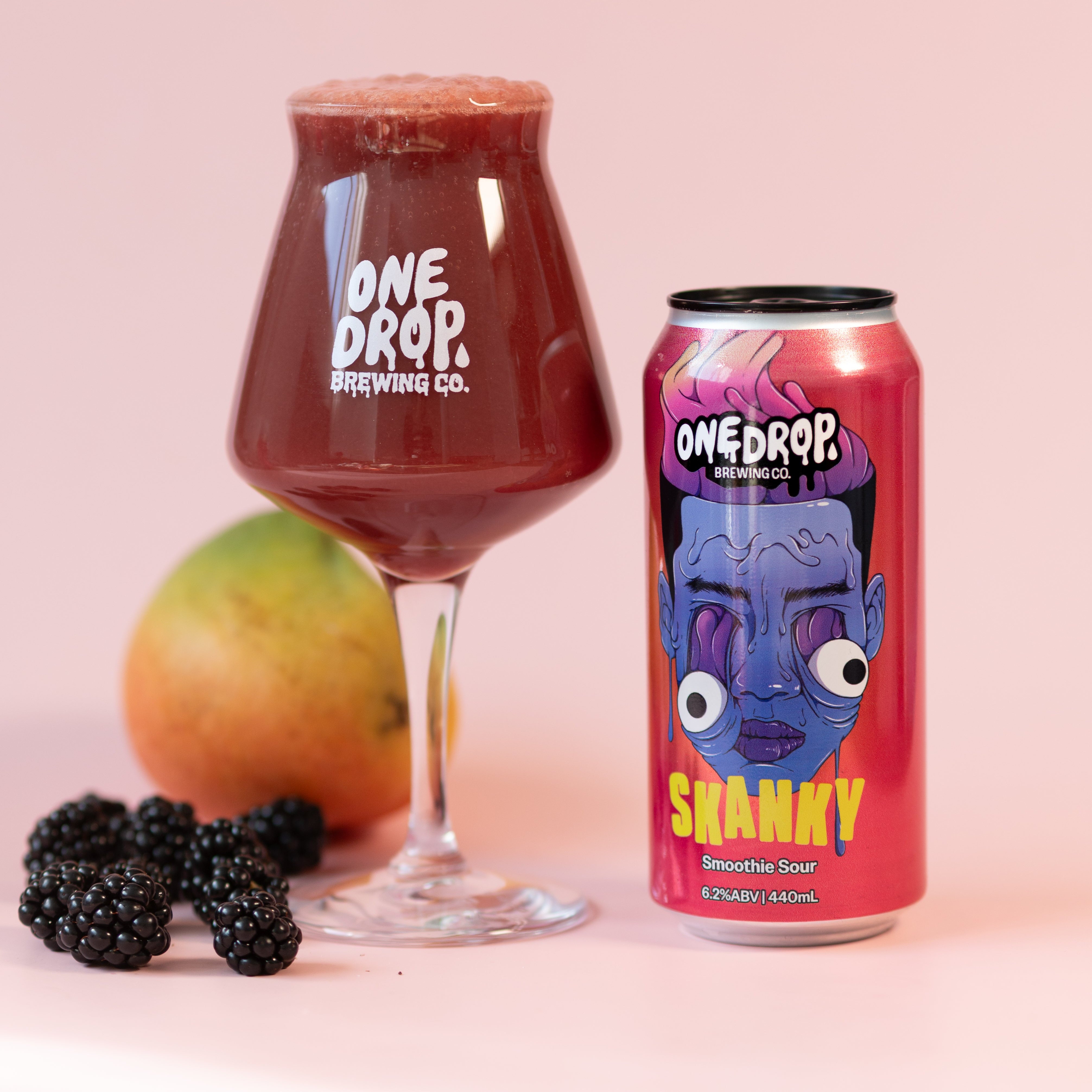 A glass of dark red beverage and a pink can of One Drop Brewing Co. Shanky sour beer, styled with a mango and blackberries on a solid light pink background.