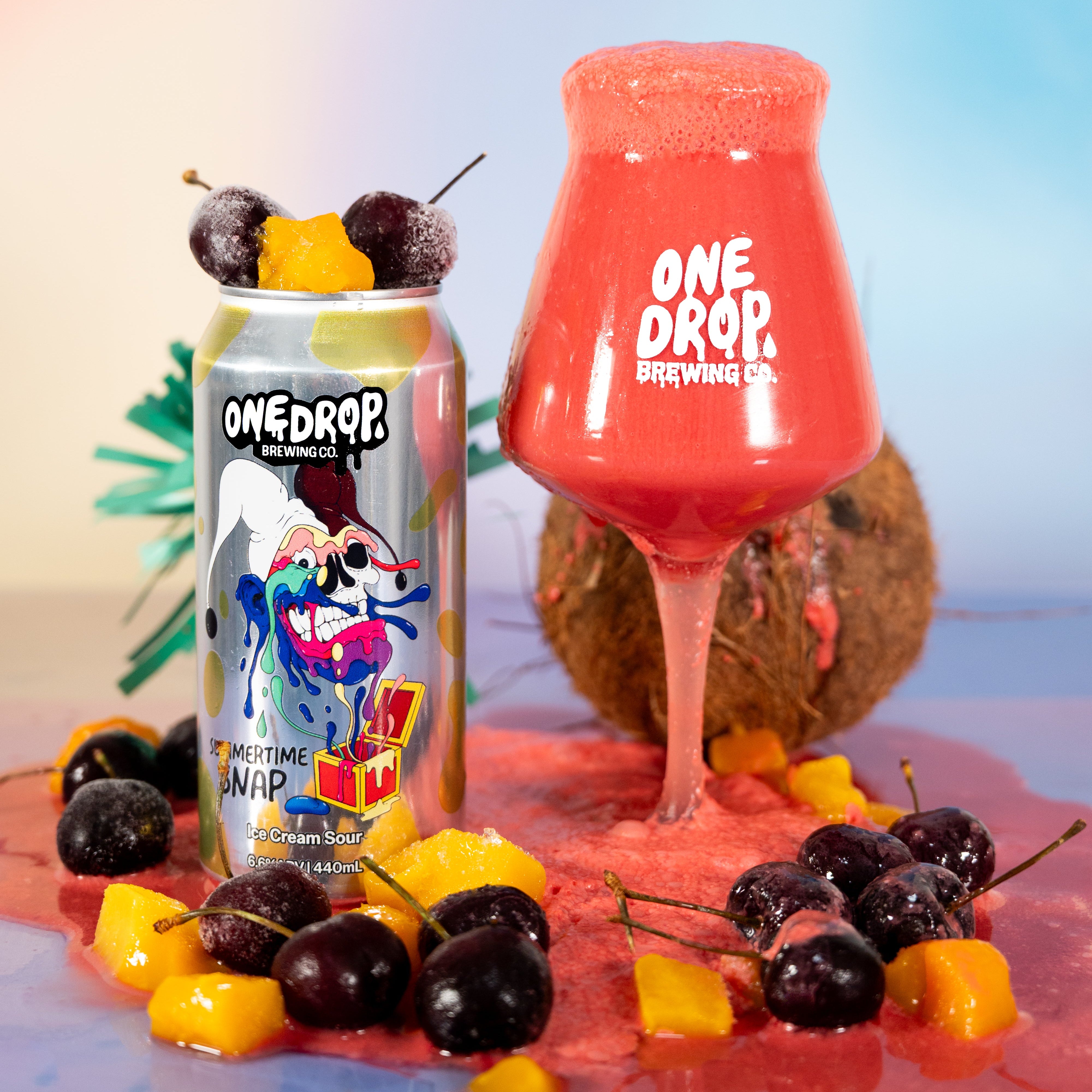 A One Drop Botanical Co. can stands next to a large glass filled with a pink-red beverage, styled with cherries, mango pieces, and a coconut on a colorful pastel background.
