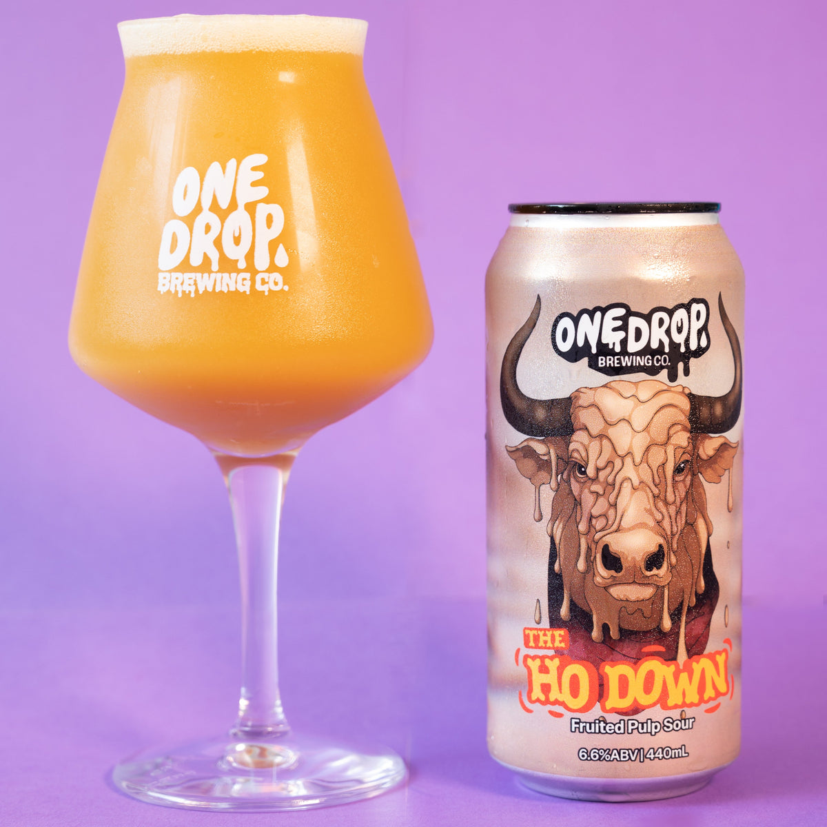 A clear stemmed glass filled with an orange-amber beer and a matching One Drop Brewing Co. can labeled 'The Ho Down', set against a solid purple background.
