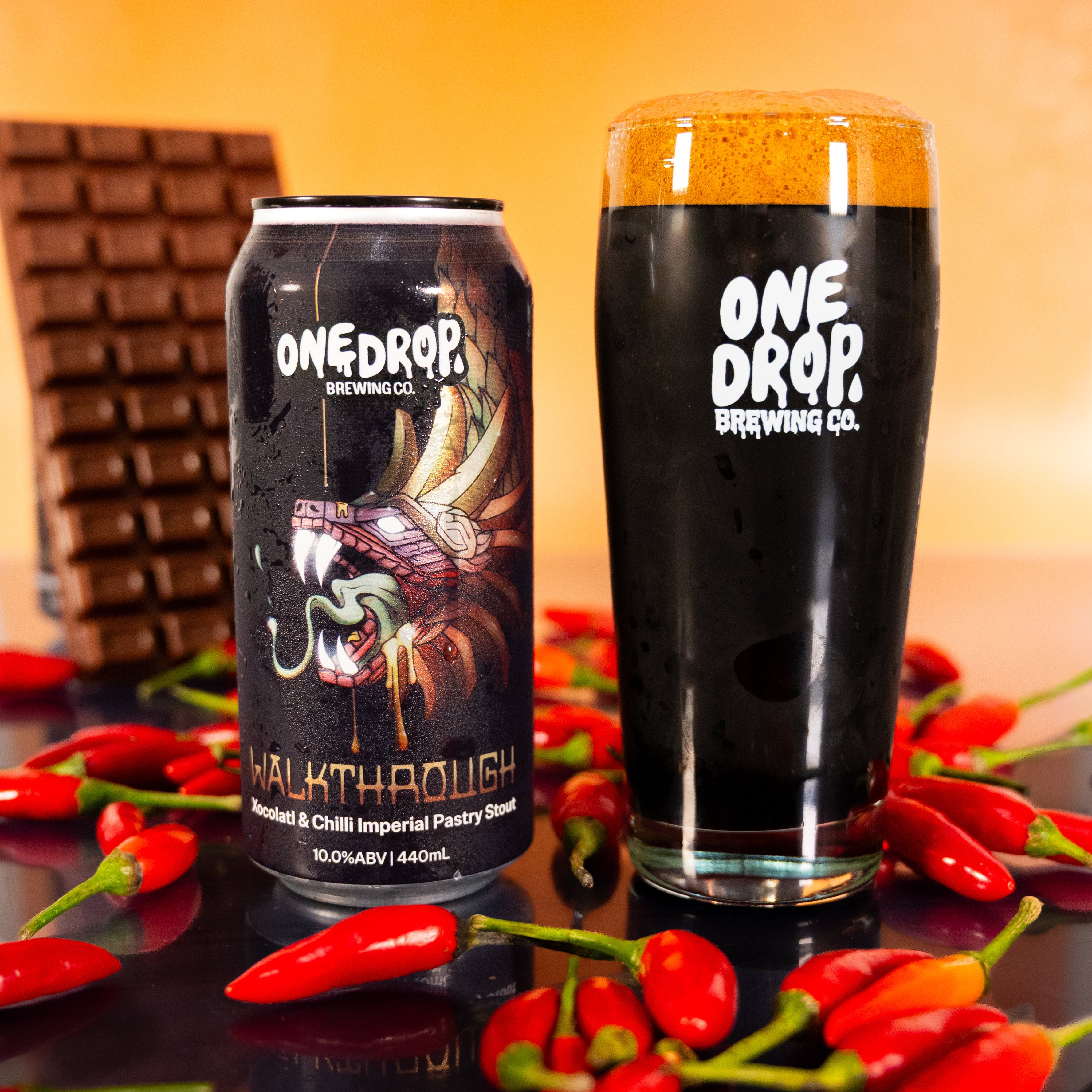 A can and a tall glass of One Drop Brewing Co. Walkthrough stout, styled with red chili peppers and a chocolate bar against a warm orange background.