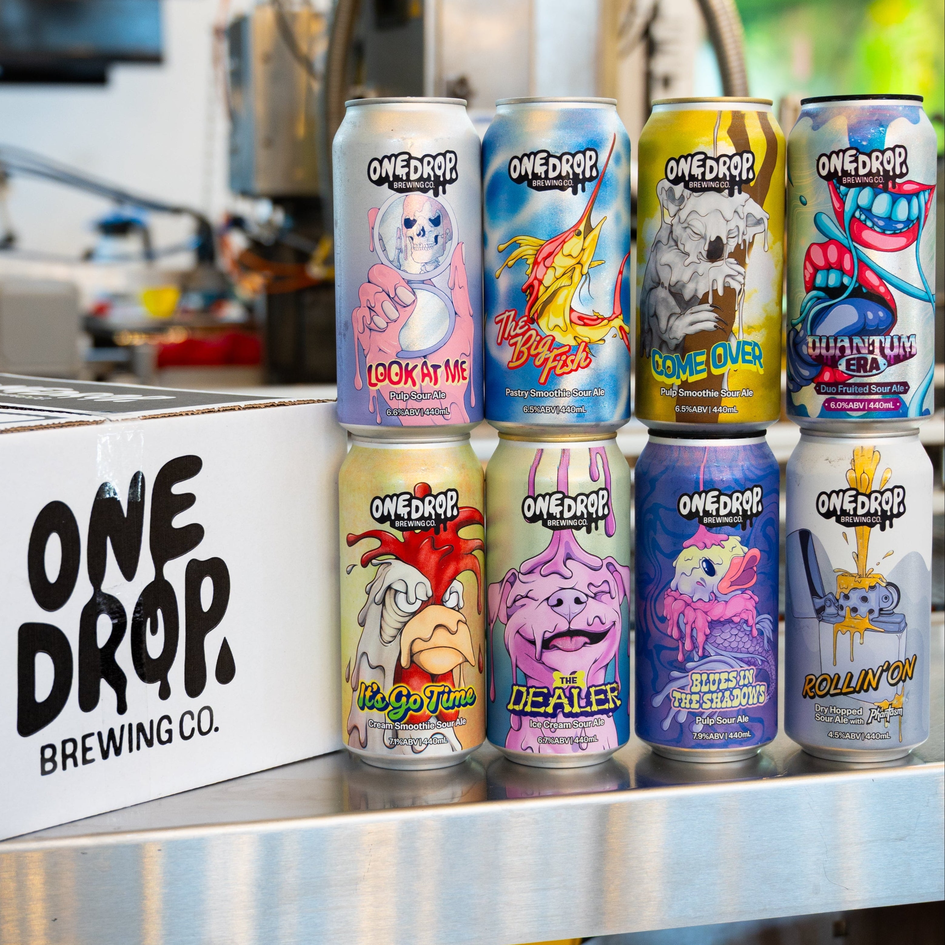 Assorted cans from One Drop Brewing Co. on a metal surface with a blurred background.
