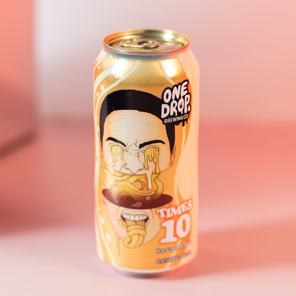 A single can of One Drop Beverages 'Times 10' dessert drink, featuring a cartoon illustration of a character with a black hairpiece and yellow face paint, set against a soft pink and peach studio background.