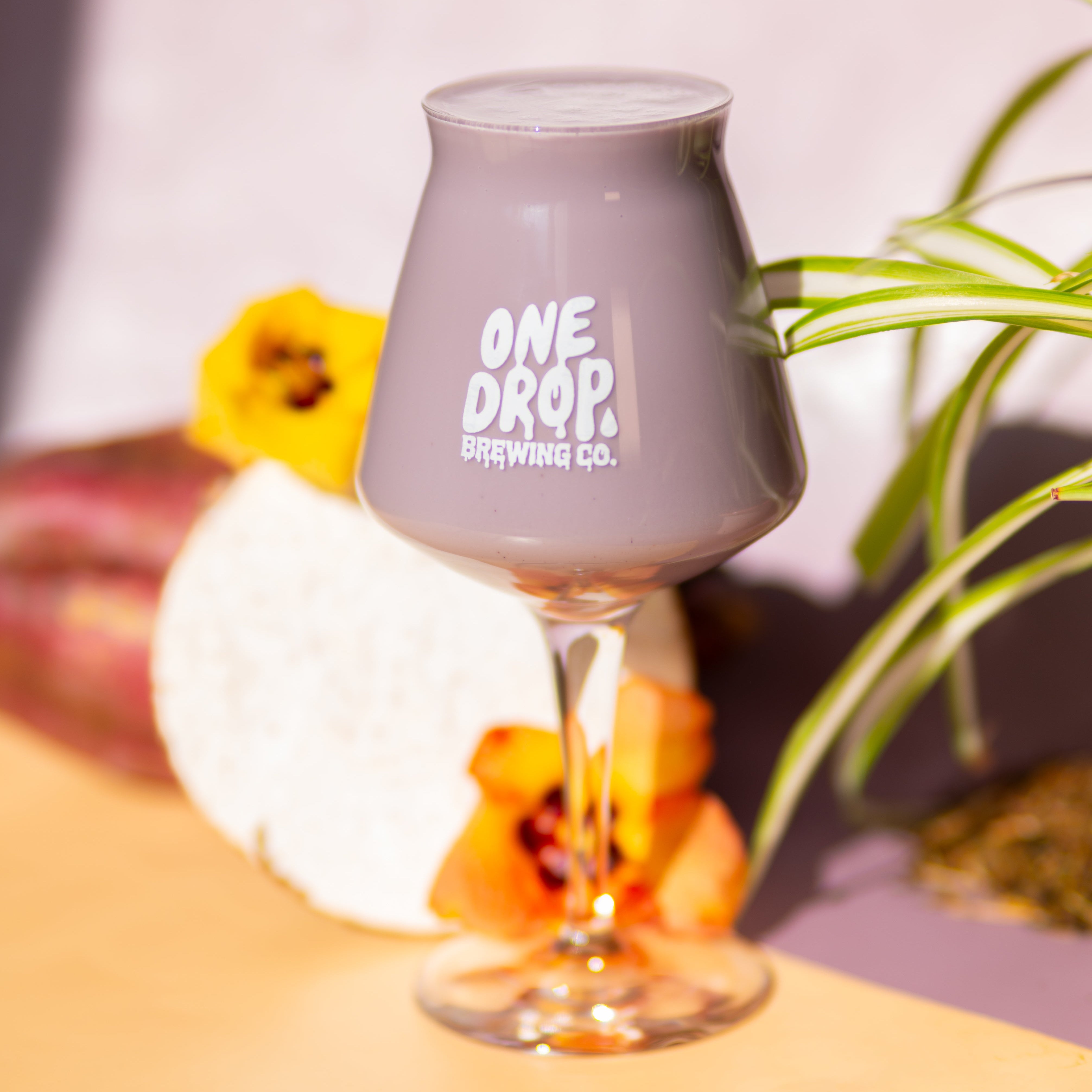 A clear stemmed glass with a gray tint and white printed text reading 'ONE DROP BREWING CO.' sits on a yellow surface, styled with flowers and green leaves against a soft lavender background.