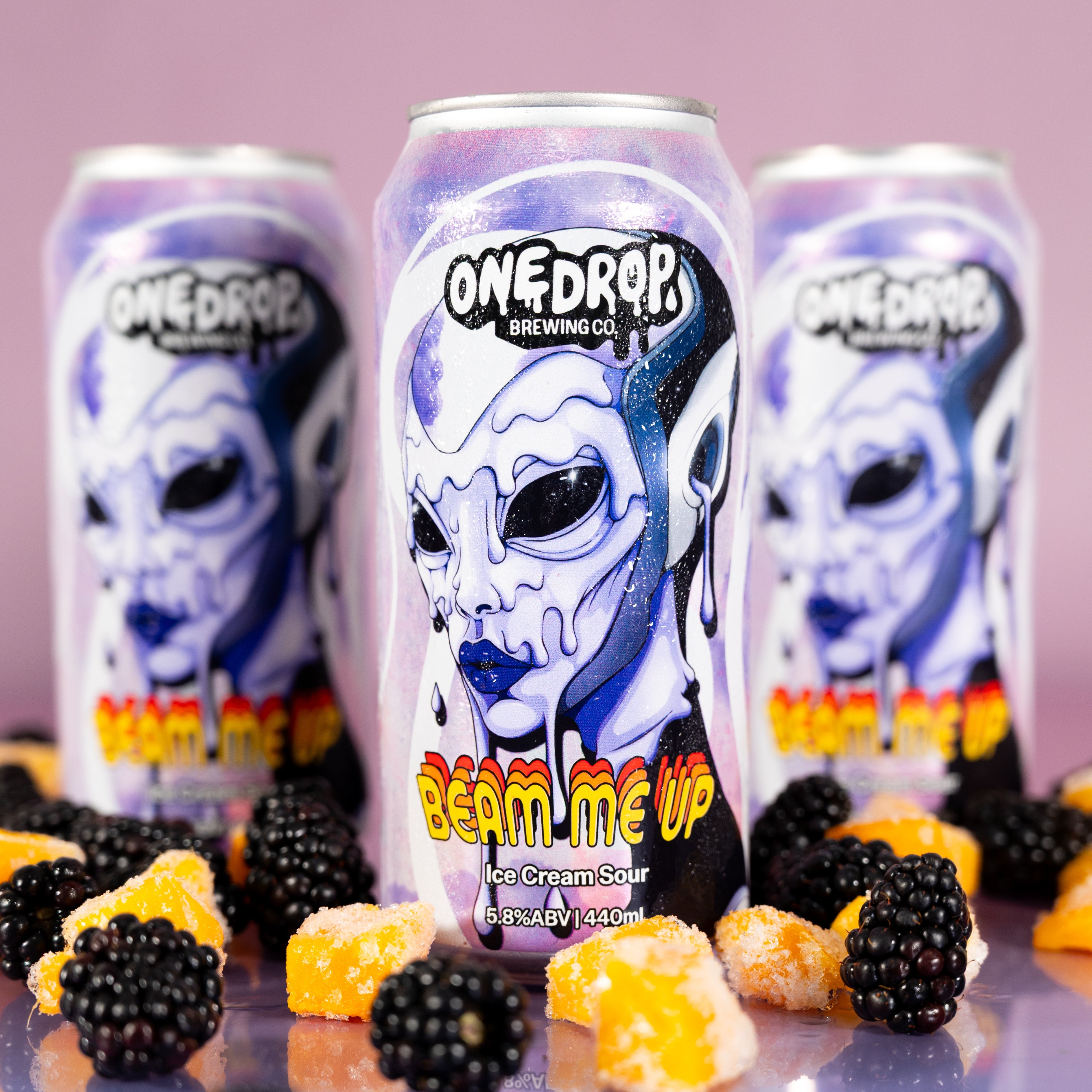 Three One Drop Brewing Co. cans of Beam Me Up ice cream sour are arranged on a reflective surface with blackberries and mango pieces in the foreground, against a pink studio background.