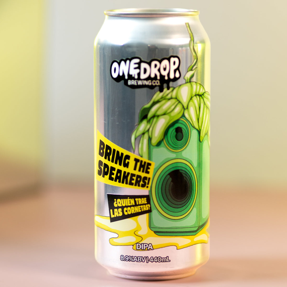 A 16 oz aluminum can of OneDrop Brewing Co. 'Bring the Speakers!' Golden Hops Lager, featuring a green hop illustration and yellow text, standing on a pink surface with a blurred beige background.