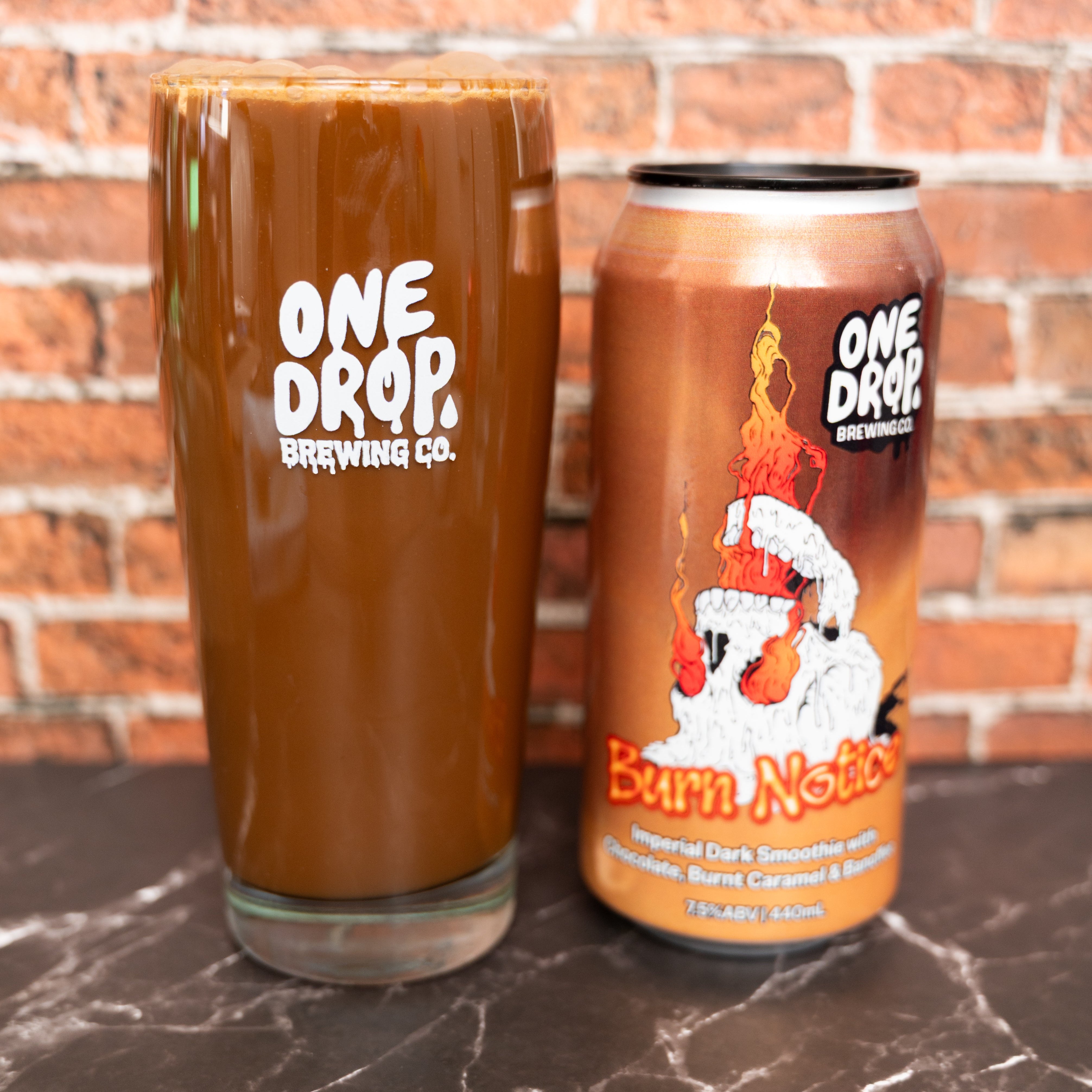 A glass of burn notice can from One Drop Brewing Co. on a dark marble surface with a brick wall background.