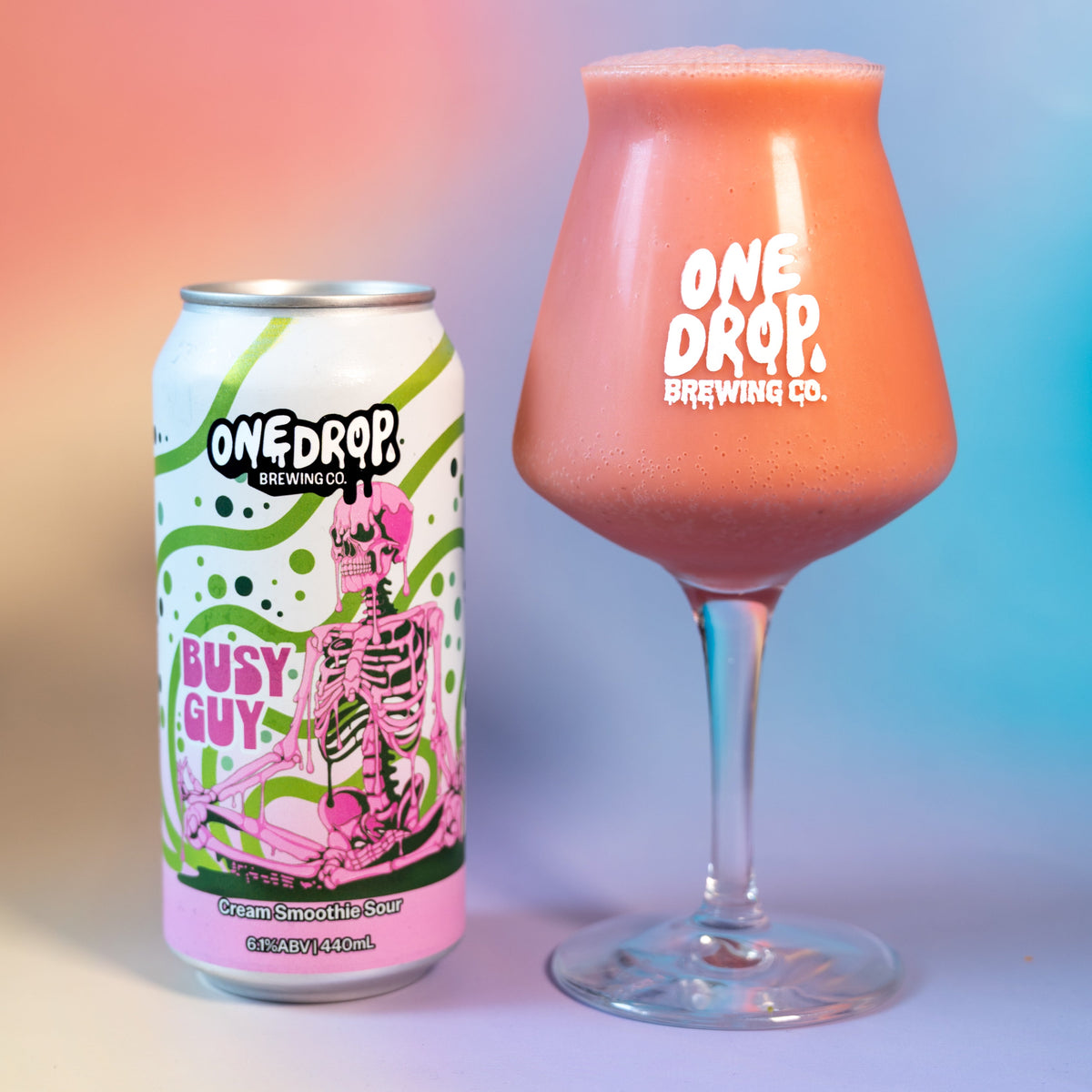 A can and a stemmed glass of One Drop Brewing Co. 'Busy Guy' beer, showing the pink and green label on the can and the orange-pink beverage inside the glass, set against a colorful pink and blue gradient background.