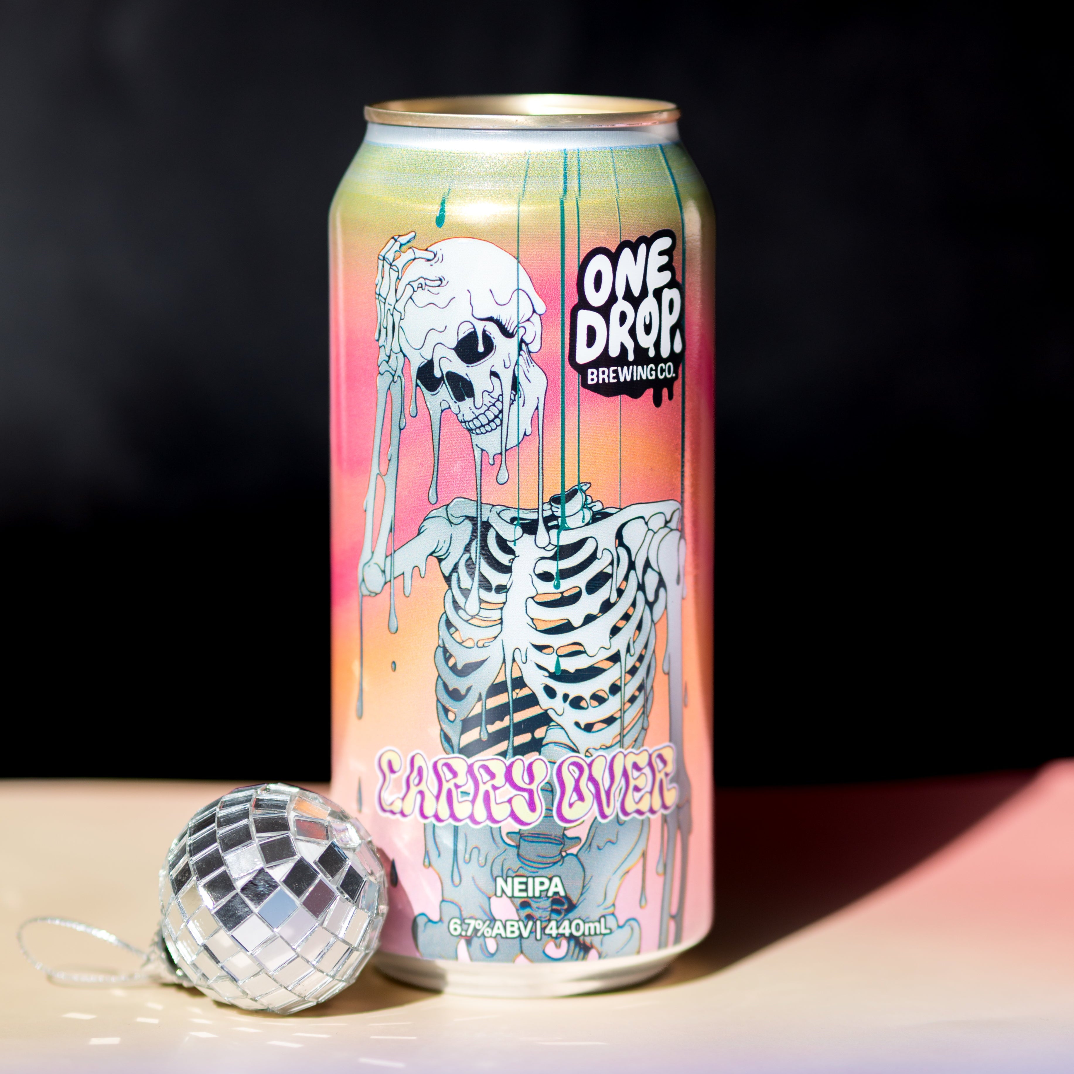 A beverage can with a colorful skeleton graphic and the text 'ONE DROP' and 'CARRY OVER' stands on a reflective surface against a dark background, with a small mirrored ball prop beside it.
