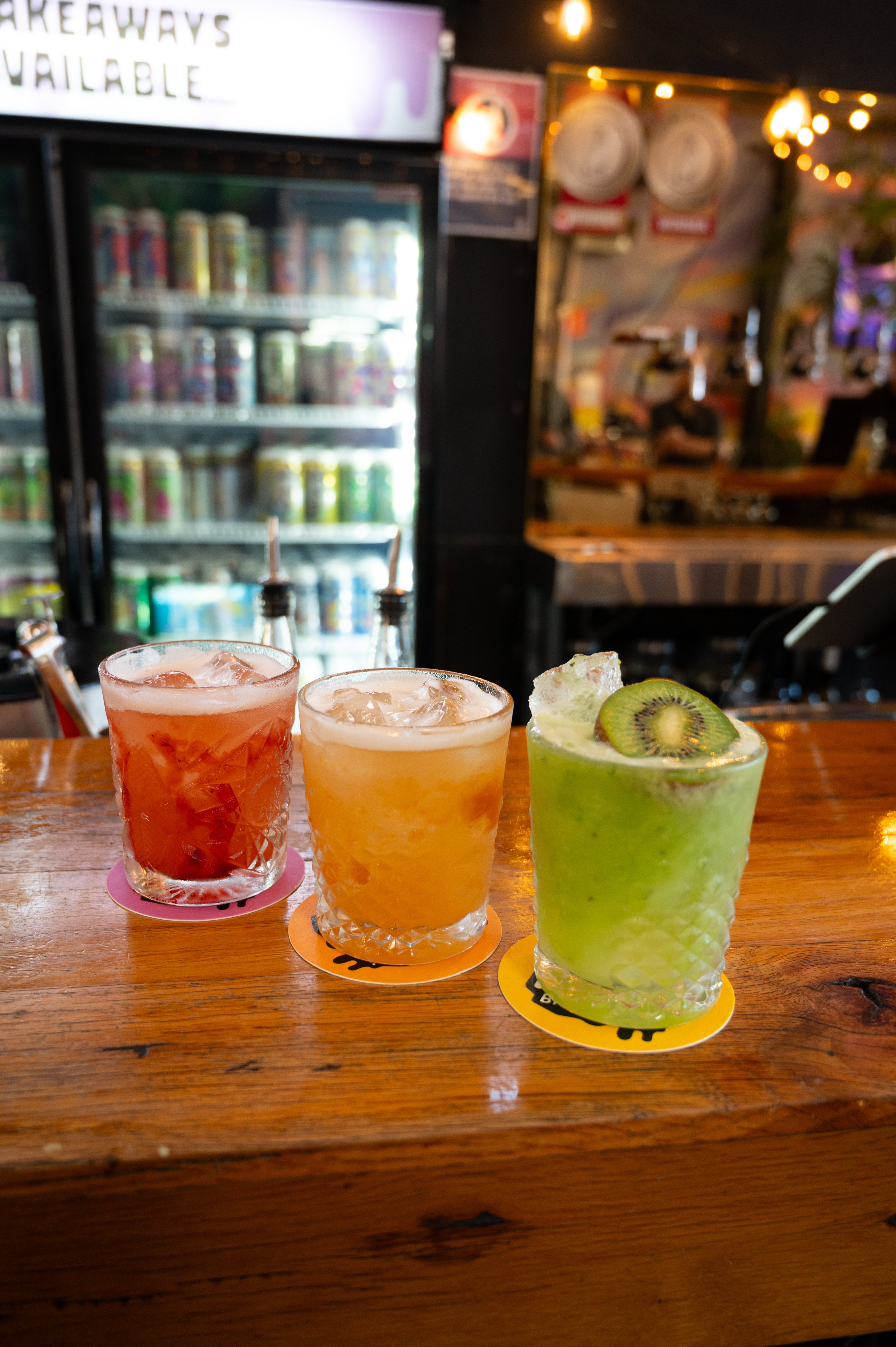 Three colorful cocktails in rocks glasses on a wooden bar counter in a bar setting. The drinks are red, orange, and green, garnished with kiwi slices, with a beverage refrigerator and bar background visible.