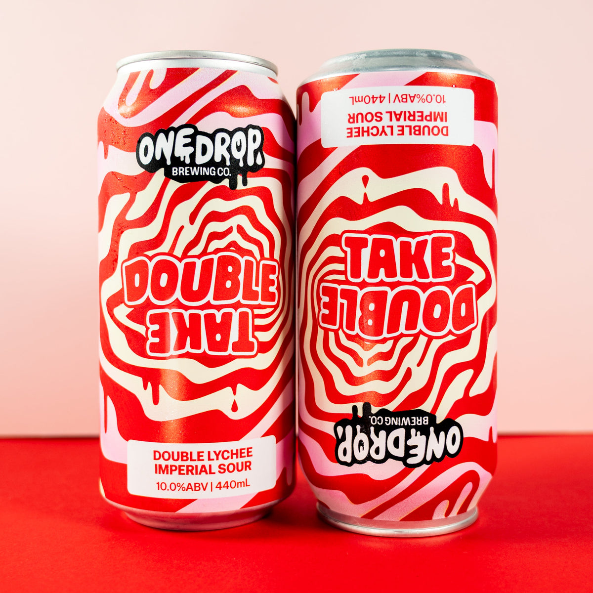 Two One Drop Beer Pongers cans of Double Lychee Imperial Sour on a red surface with a pale pink background.