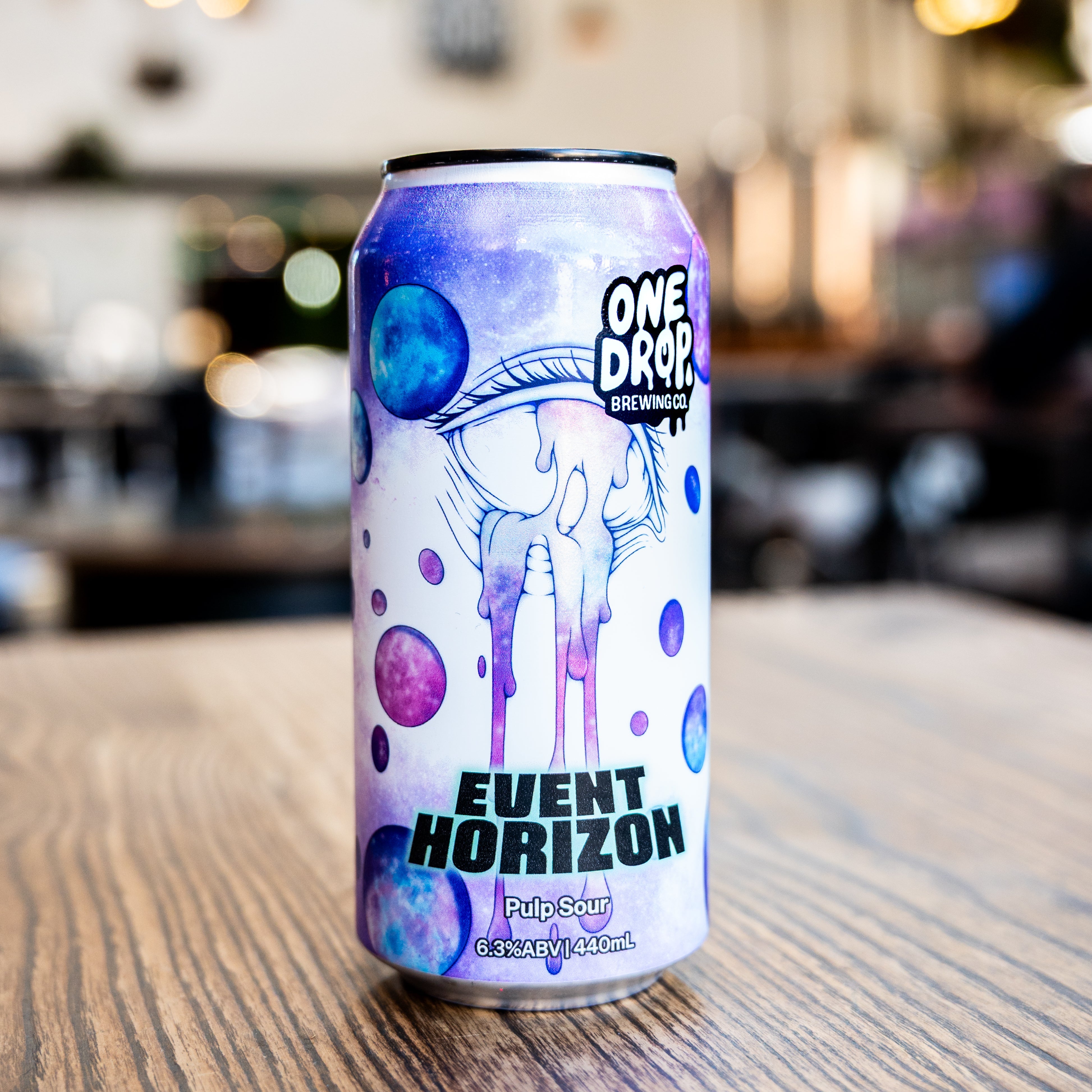 A single can of One Drop Brewing Event Horizon Pilsner Lager sits on a wooden table in a blurred indoor bar or cafe setting.
