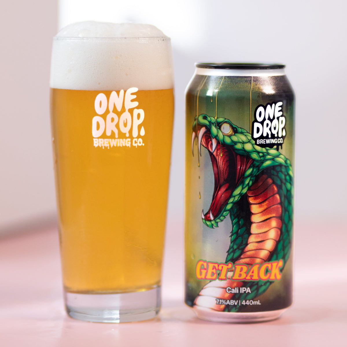 A glass of golden beer with white foam and a matching One Drop Brewing Canvases 'Get Back' craft IPA can, displayed side-by-side on a pink surface against a white background.