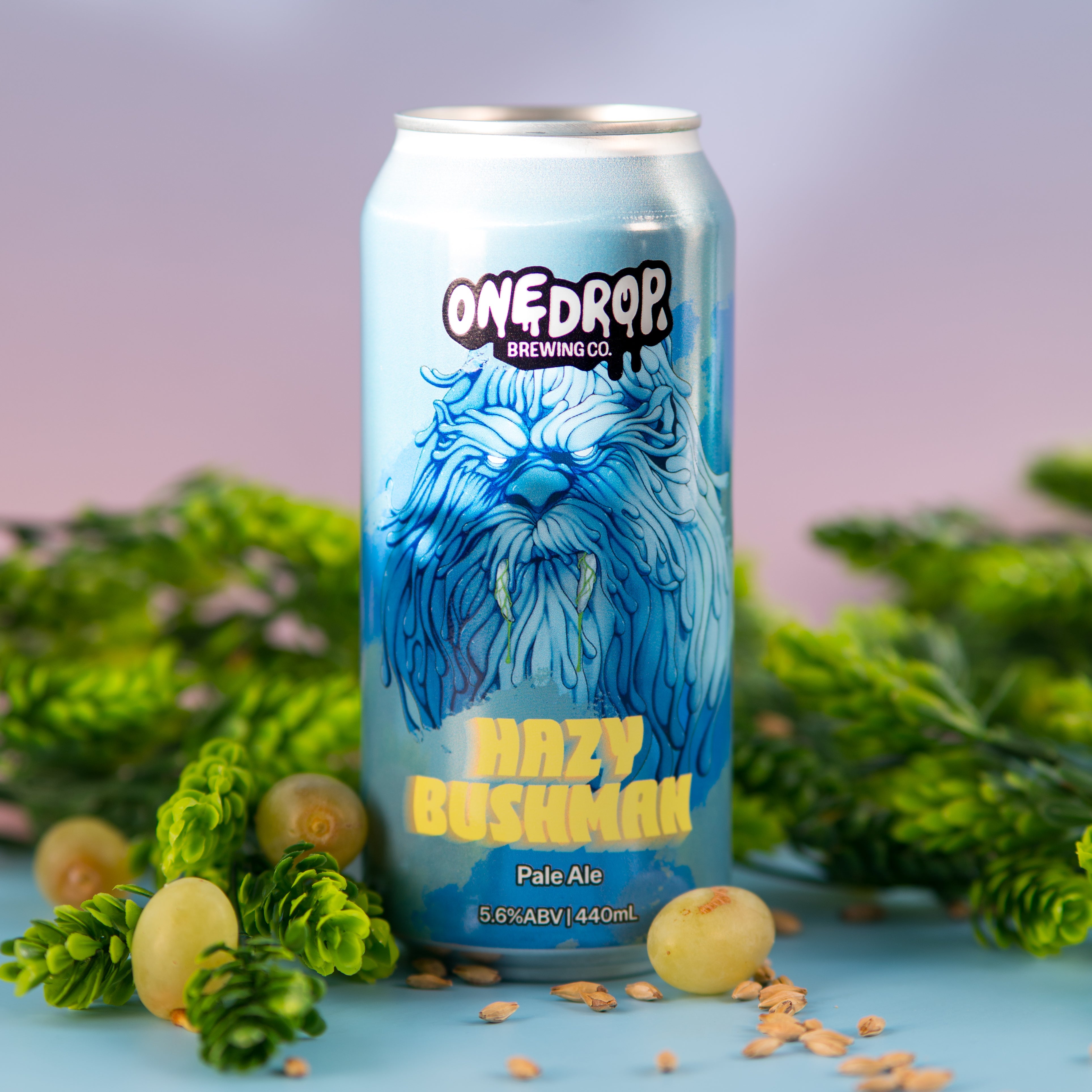 A 33cl aluminum can of OneDrop Brewing's 'Mazy Boshman' Pale Ale stands upright on a light blue surface. The can features a vibrant blue illustration of a shaggy dog and is surrounded by green pinecones and scattered malt beads, with a soft-focus background.