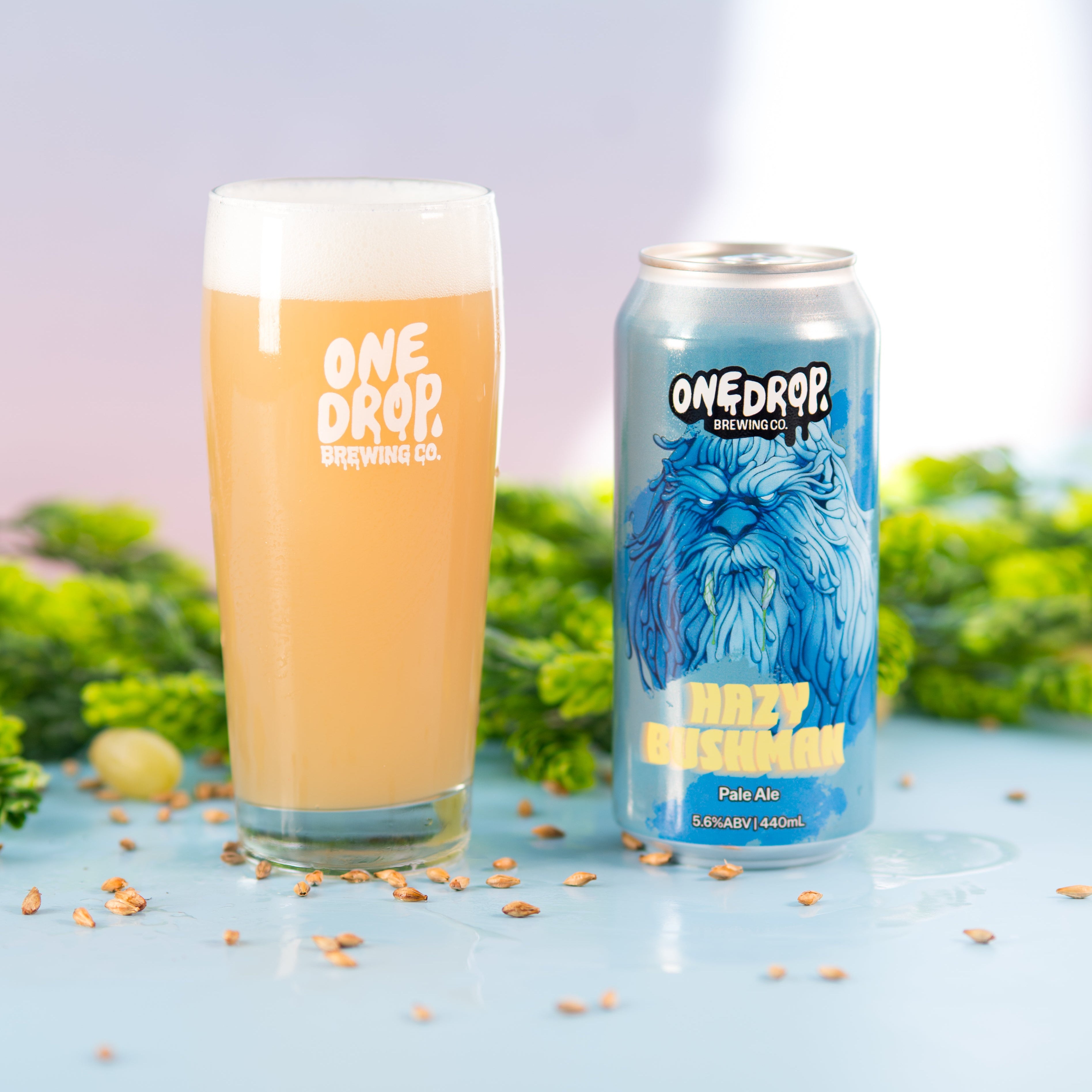 A glass of frothy pale orange beer and a blue can of One Drop Brewing Co. 'Buzzy Bummer' IPA stand on a light blue surface with green herbs and scattered hops around them.
