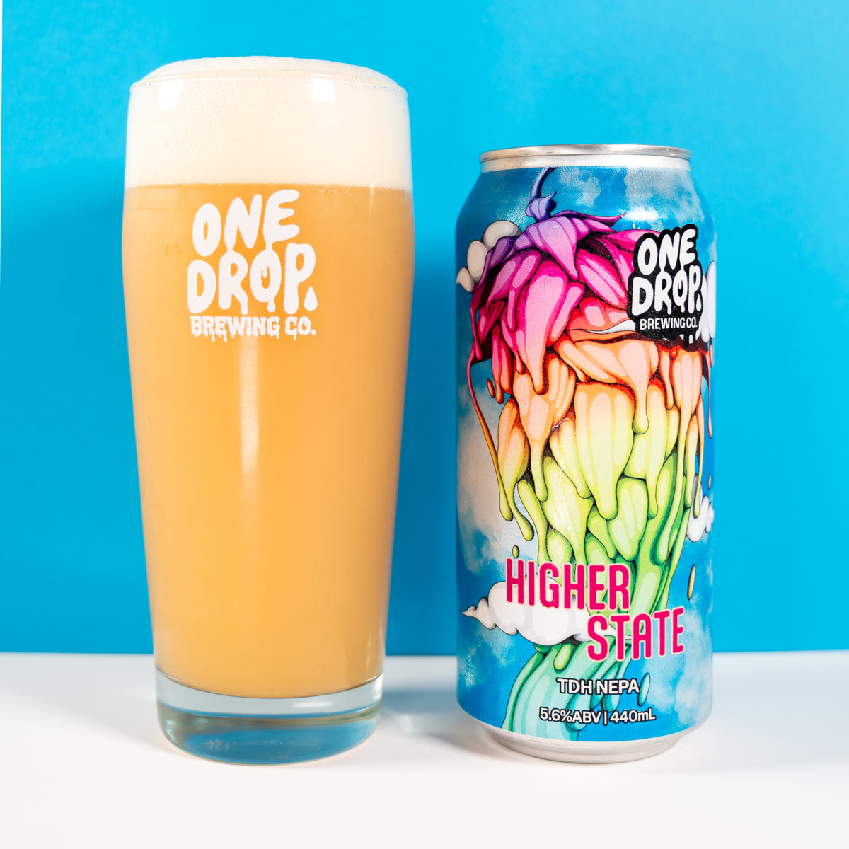 A glass of frothy golden beer and a can of One Drop Brewing Co. Higher State IPA next to each other on a white surface against a bright blue background.