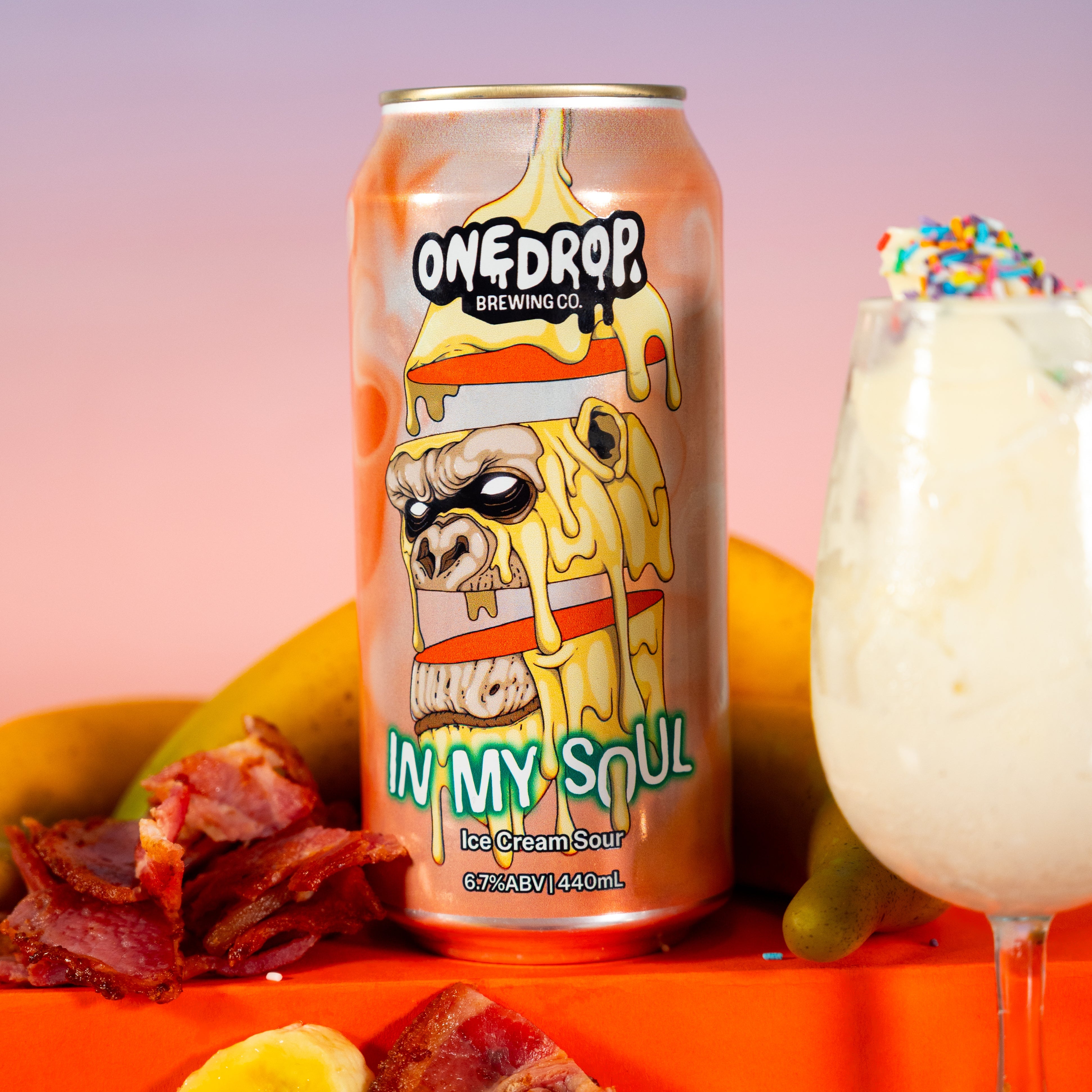 A 12 fl oz can of OneDrop Breakfasts 'In My Soul' ice cream sour beer is displayed alongside a glass of creamy beverage, bananas, bacon, and salt on an orange surface against a pink and blue gradient background.