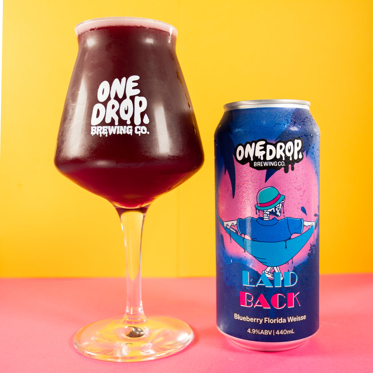 A can of One Drop Brewing Co. Laid Back Blueberry Florida Weiss alongside a large tulip glass filled with the dark purple beverage, set against a bright yellow background and pink surface.