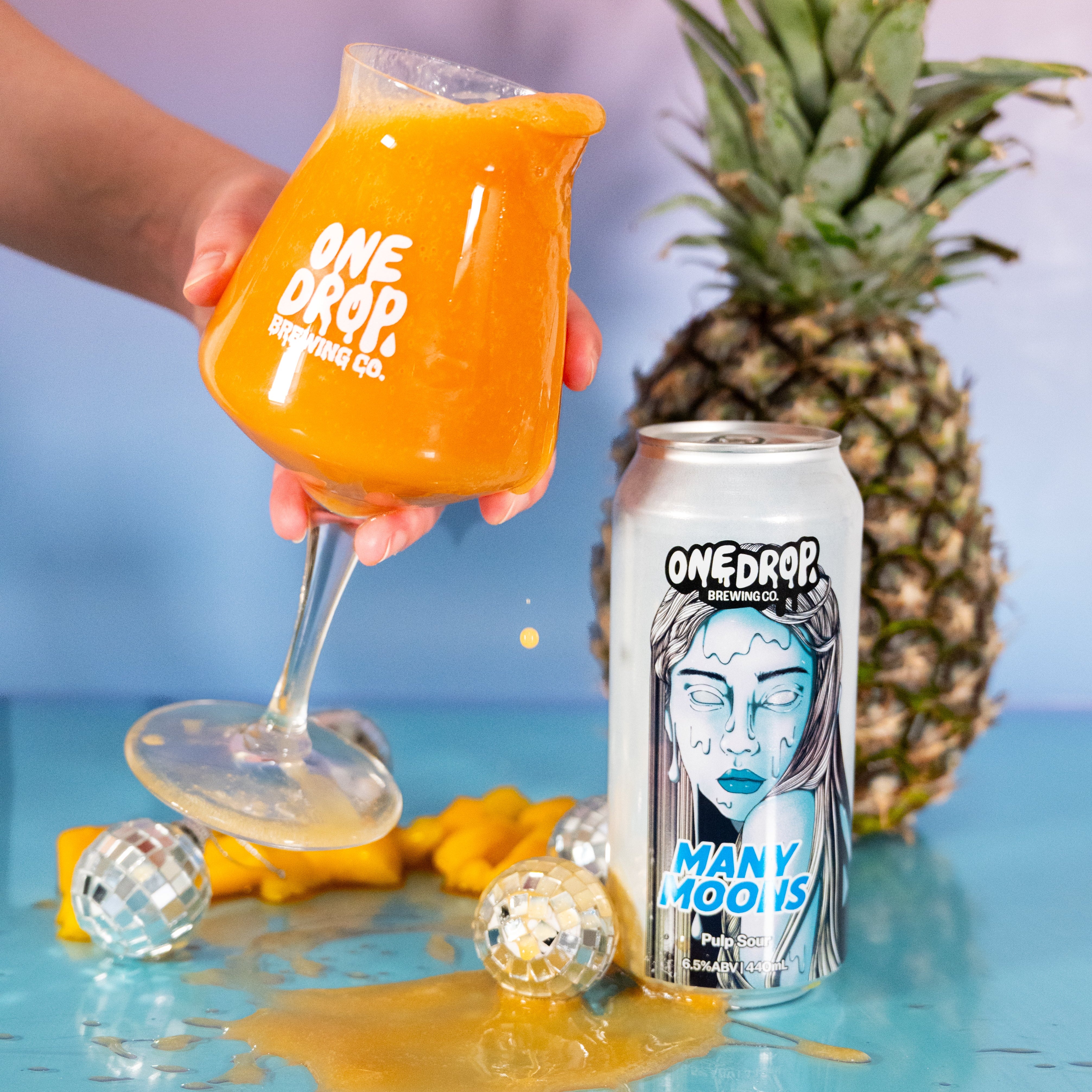 A hand holds a stemmed glass filled with bright orange drink above a spilled puddle, next to a One Drop Brewing Co. can labeled 'Many Moons' and a whole pineapple on a blue surface.