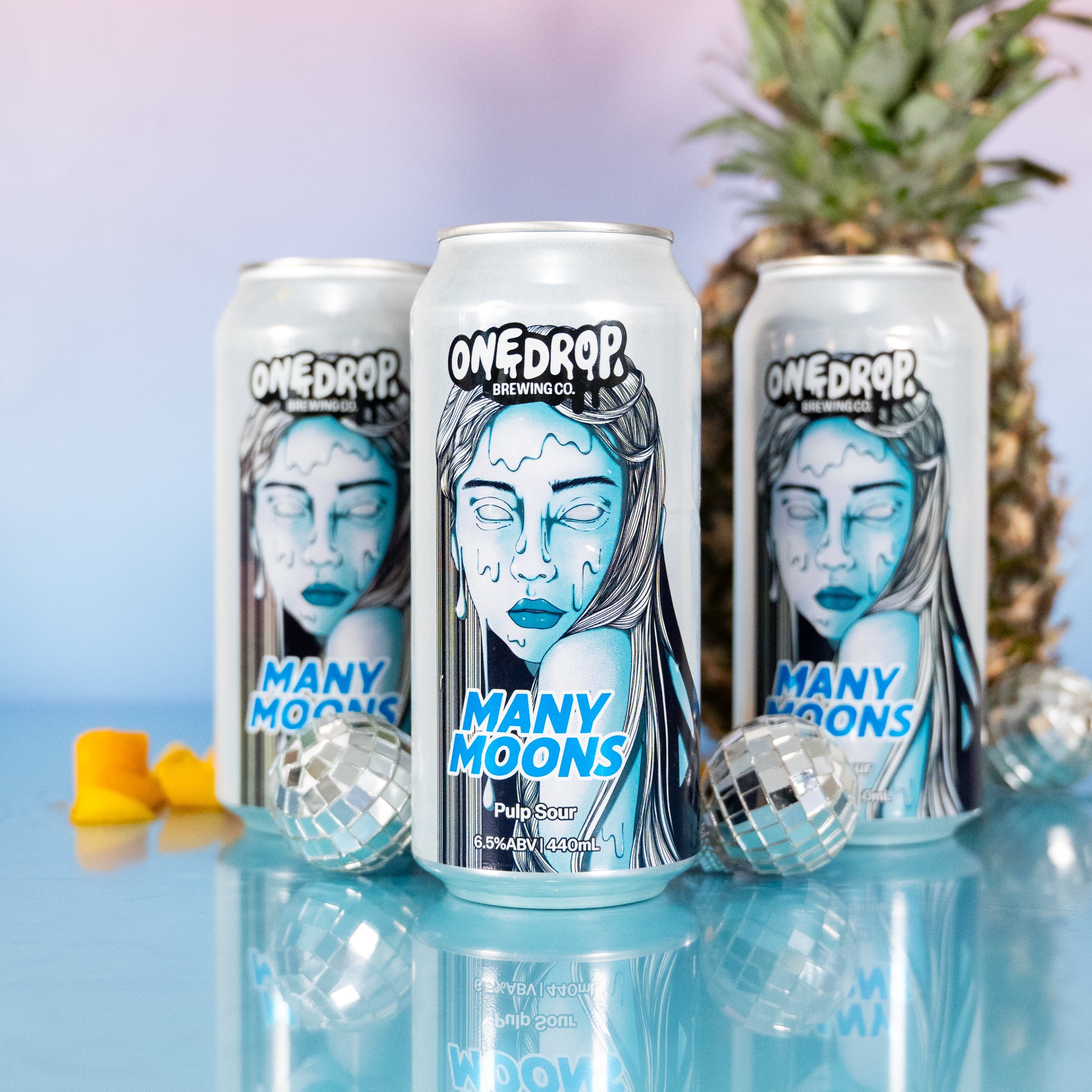 Three OneDrop 'Many Moons' beverage cans arranged on a reflective light blue surface, with a pineapple and small yellow candies in the background against a pink-to-blue gradient backdrop.