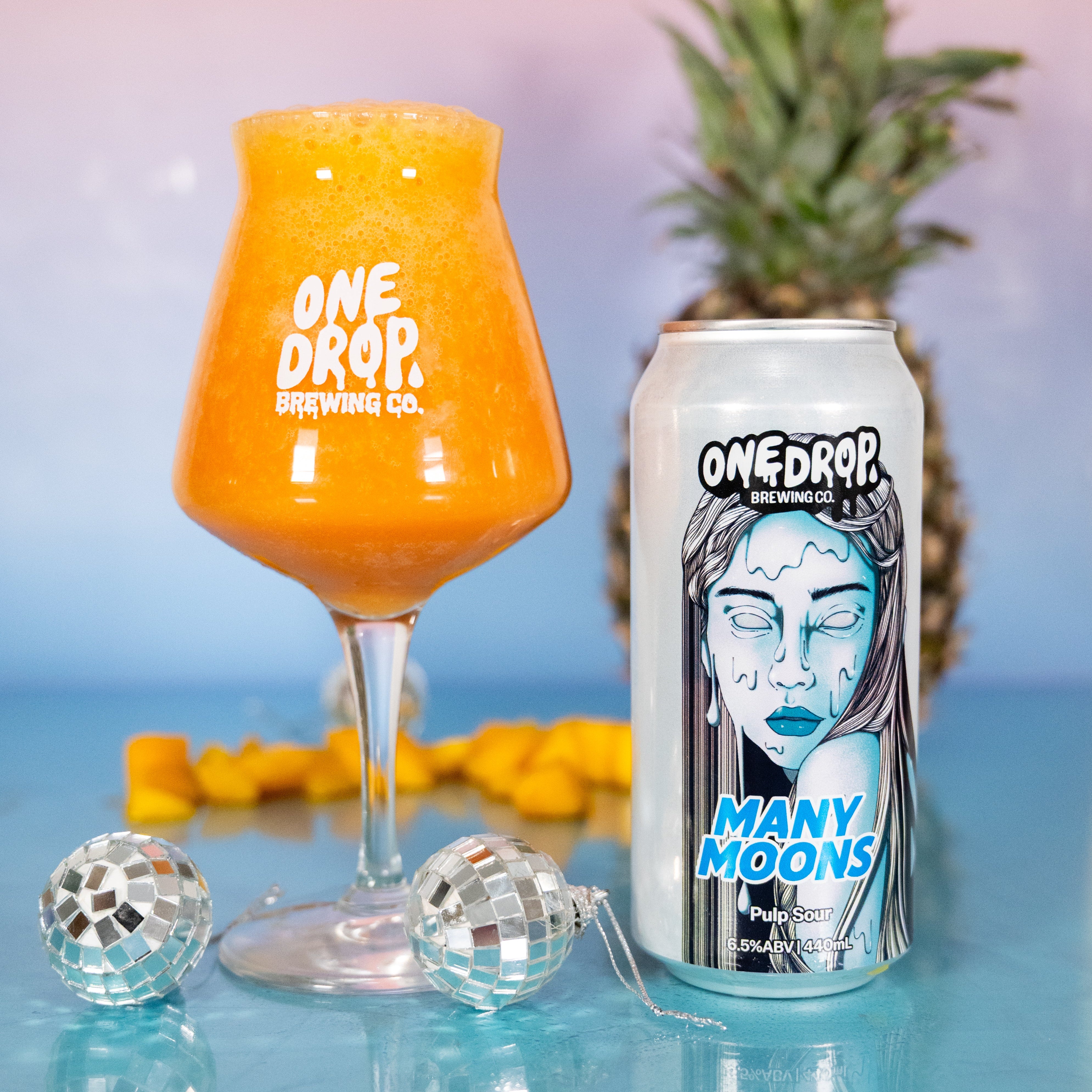 A One Drop Brewing Co. can of 'Many Moons' next to a stemmed glass filled with an orange drink, styled with a pineapple, disco balls, and yellow fruit on a reflective blue surface.