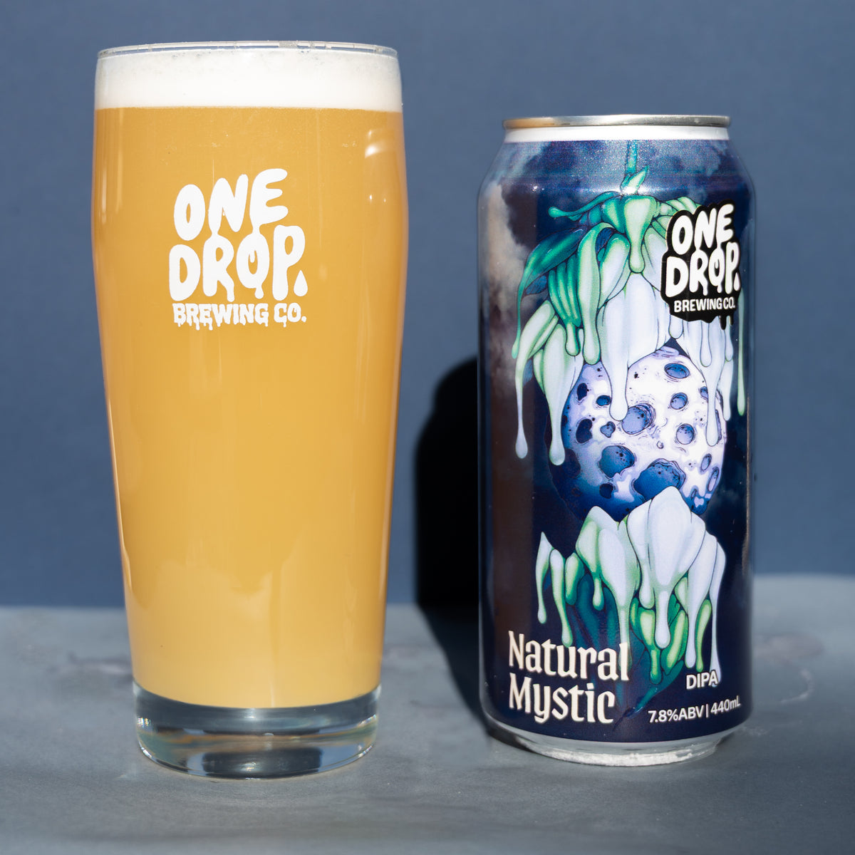 A pint glass filled with amber beer and a can of One Drop Brewing Co. Natural Mystic DIPA standing beside it on a gray surface against a blue-gray background.