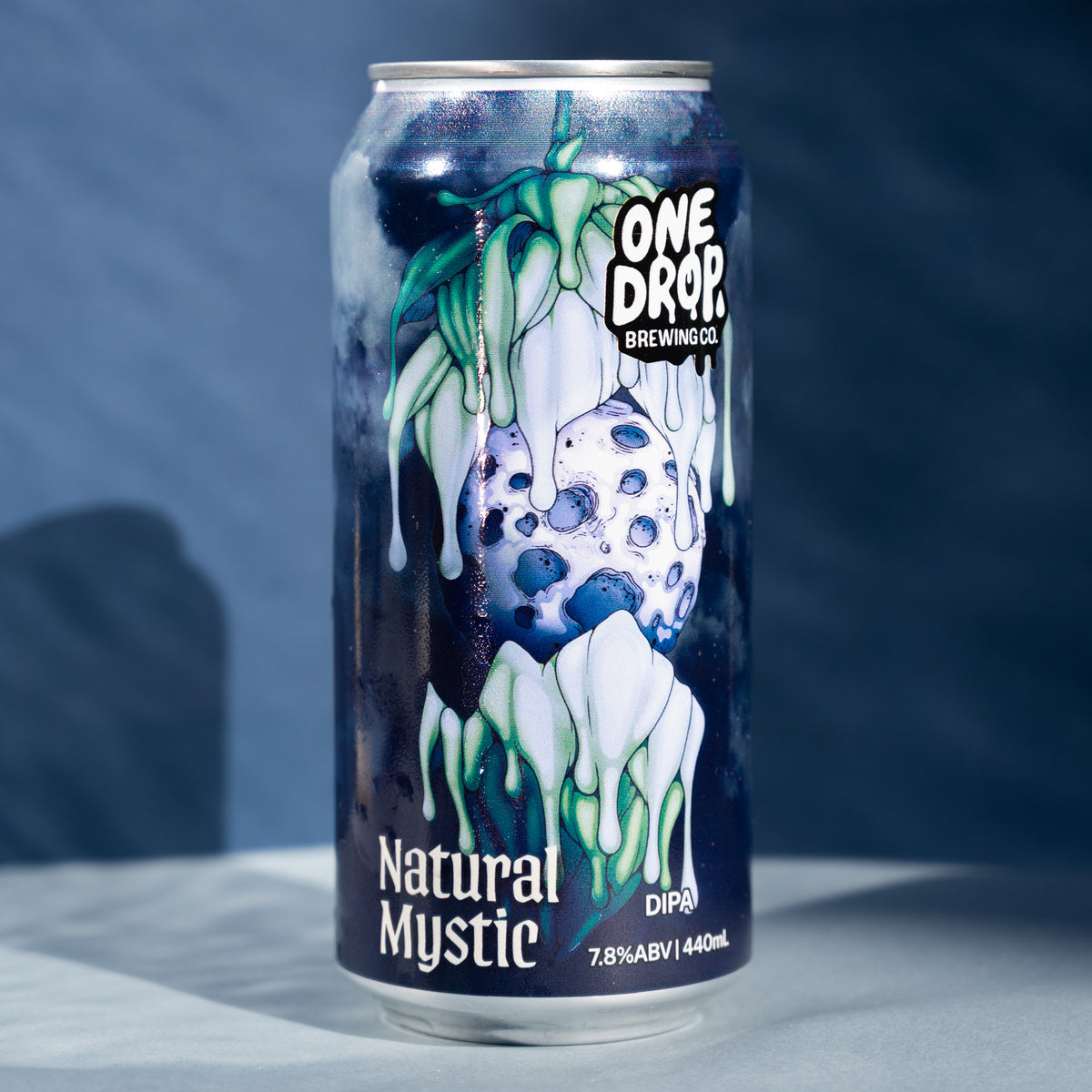 A 440mL aluminum can of One Drop She Winkool 'Natural Mystic' beverage, featuring a dark blue and white abstract dripping design with green accents, set against a dark blue textured background.
