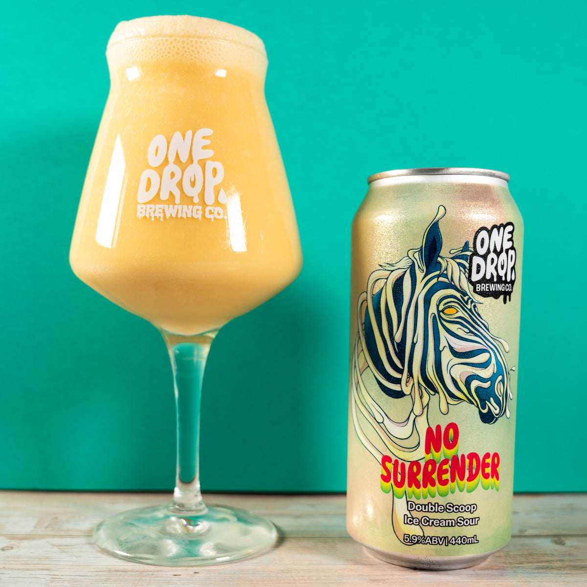 A can and a stemmed glass of One Drop Brewing Co. 'No Surrender' double scoop creamy sour apple pie beverage, shown on a light wood surface against a solid teal background.