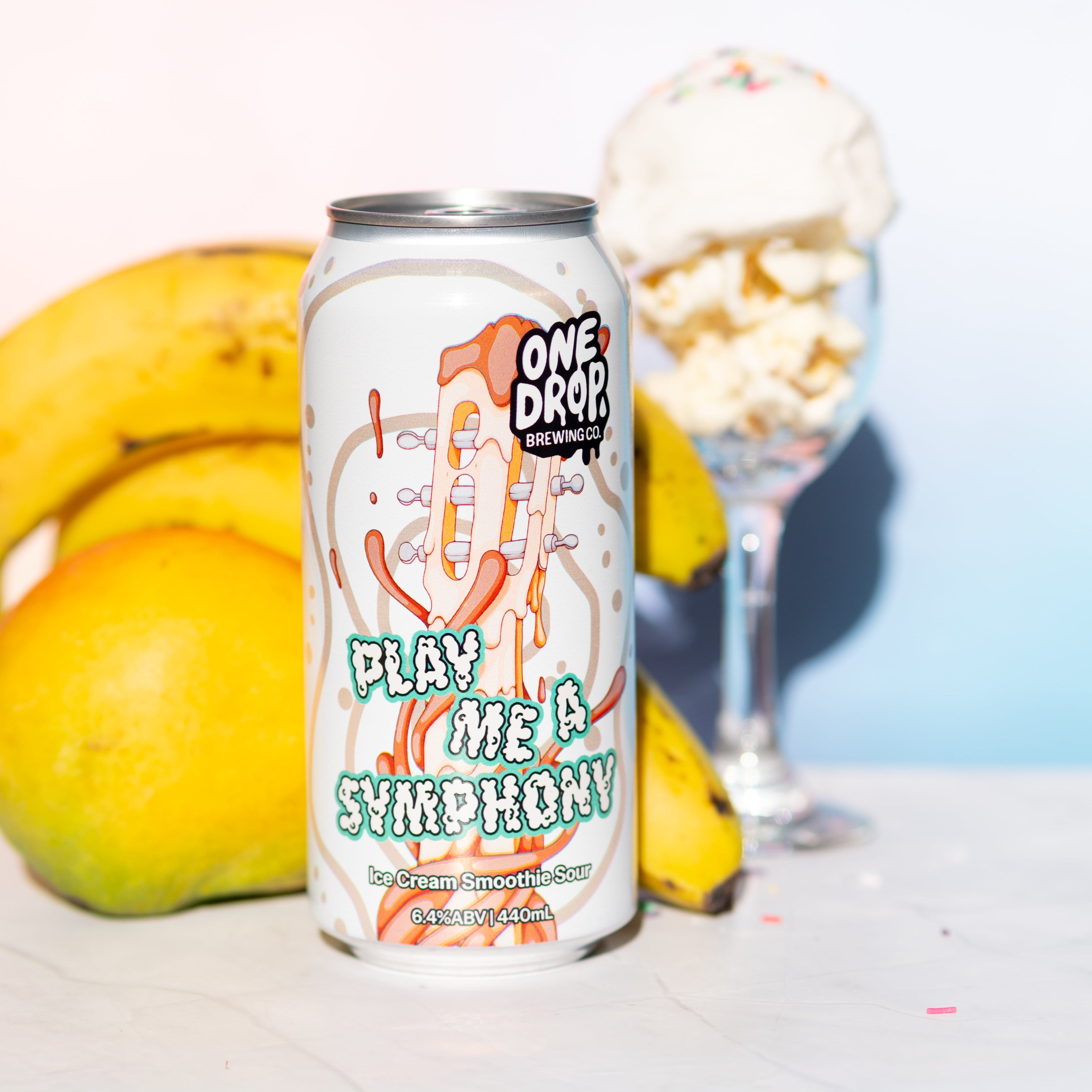 A can of One Drop Coffee Roasters 'Play Me A Symphony' cold brew coffee stands on a white surface, styled with bananas and a glass of ice cream in the background against a light blue and white gradient backdrop.