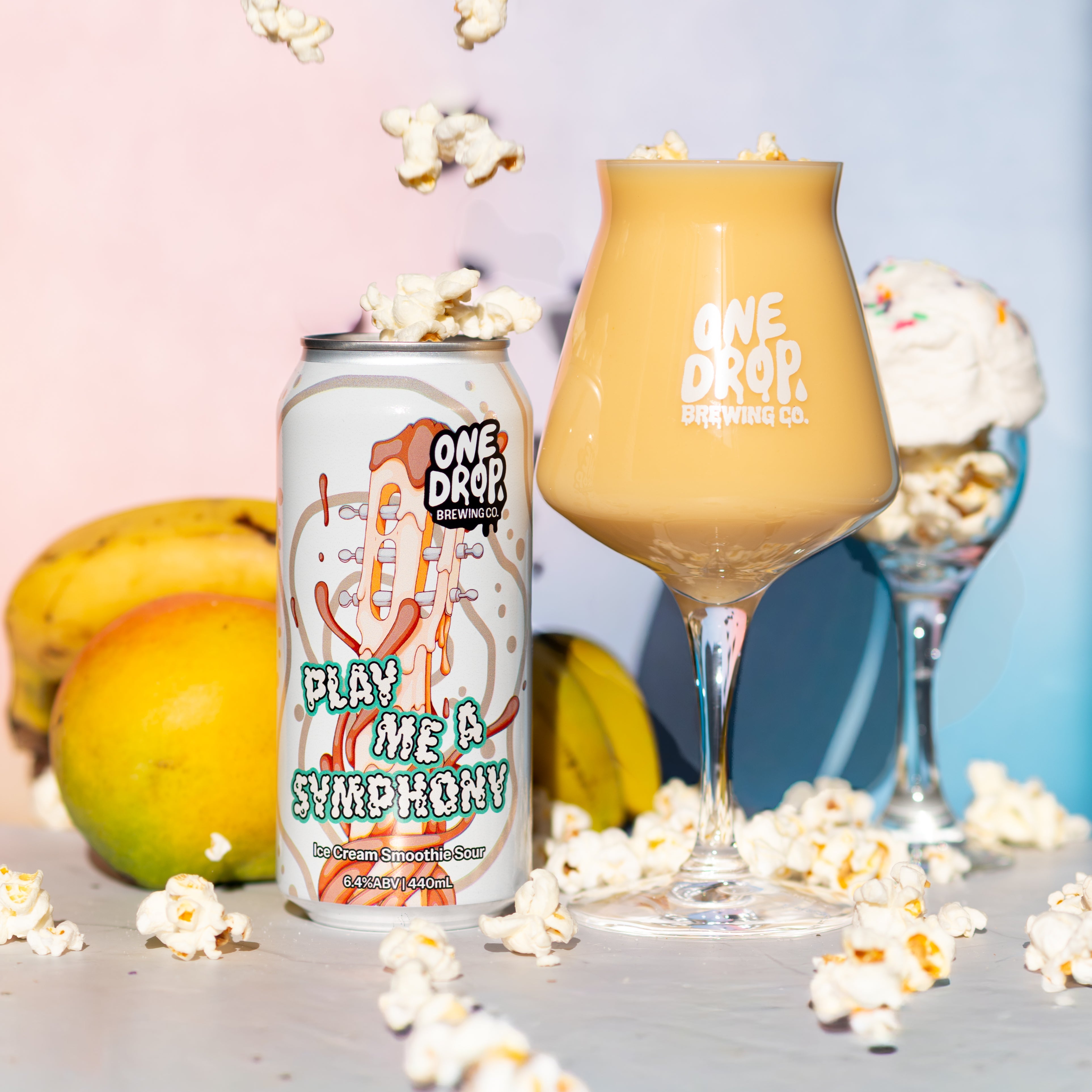 A beverage can labeled One Drop Brewing Co. next to a filled stemmed glass and a dessert glass, styled with floating popcorn and fruit on a pastel pink and blue background.
