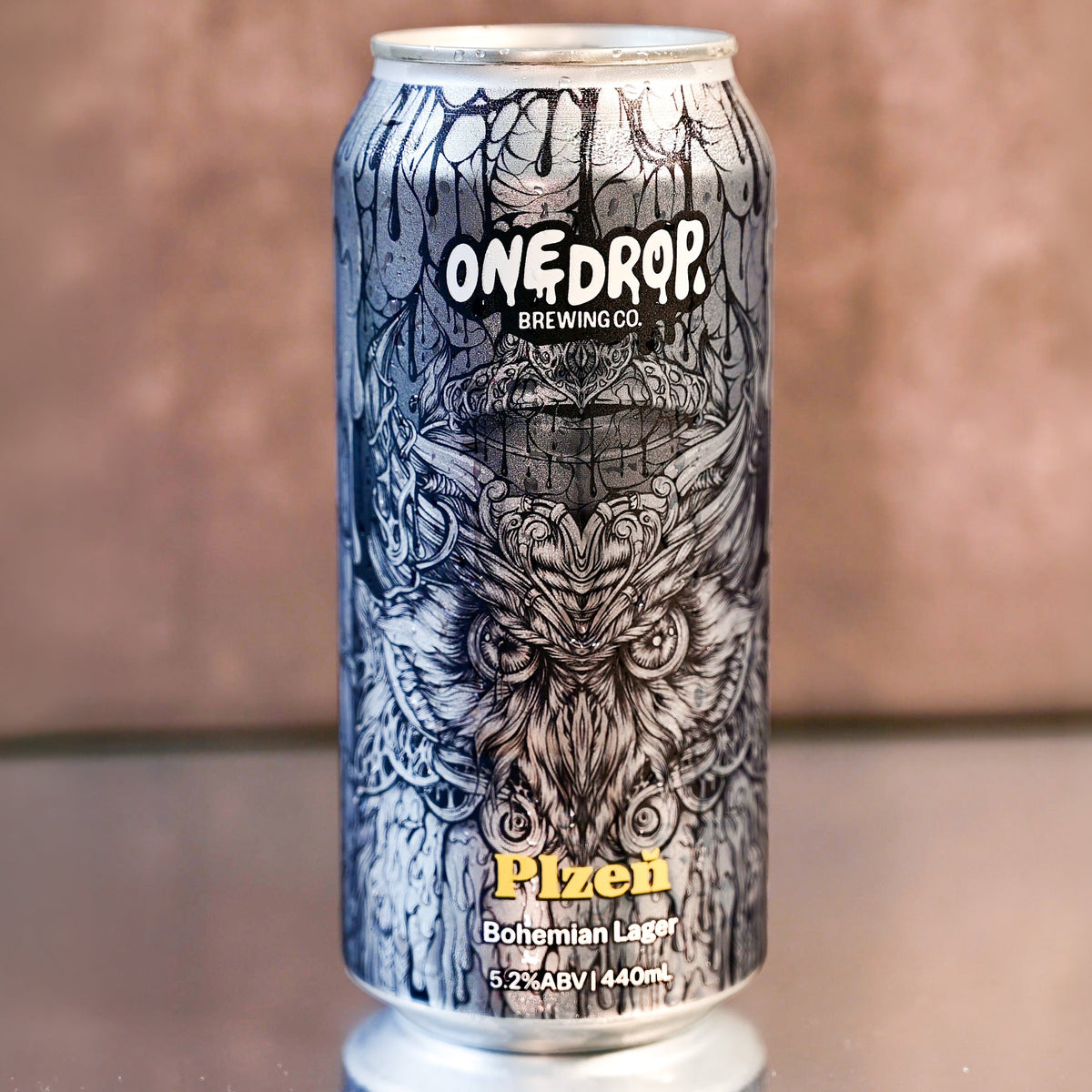 A 12 oz aluminum can of OneDrop Brewing Co. Piezêh Bohemian Lager, featuring a detailed black and white illustration of an owl on a tree trunk, set against a blurred brown background.
