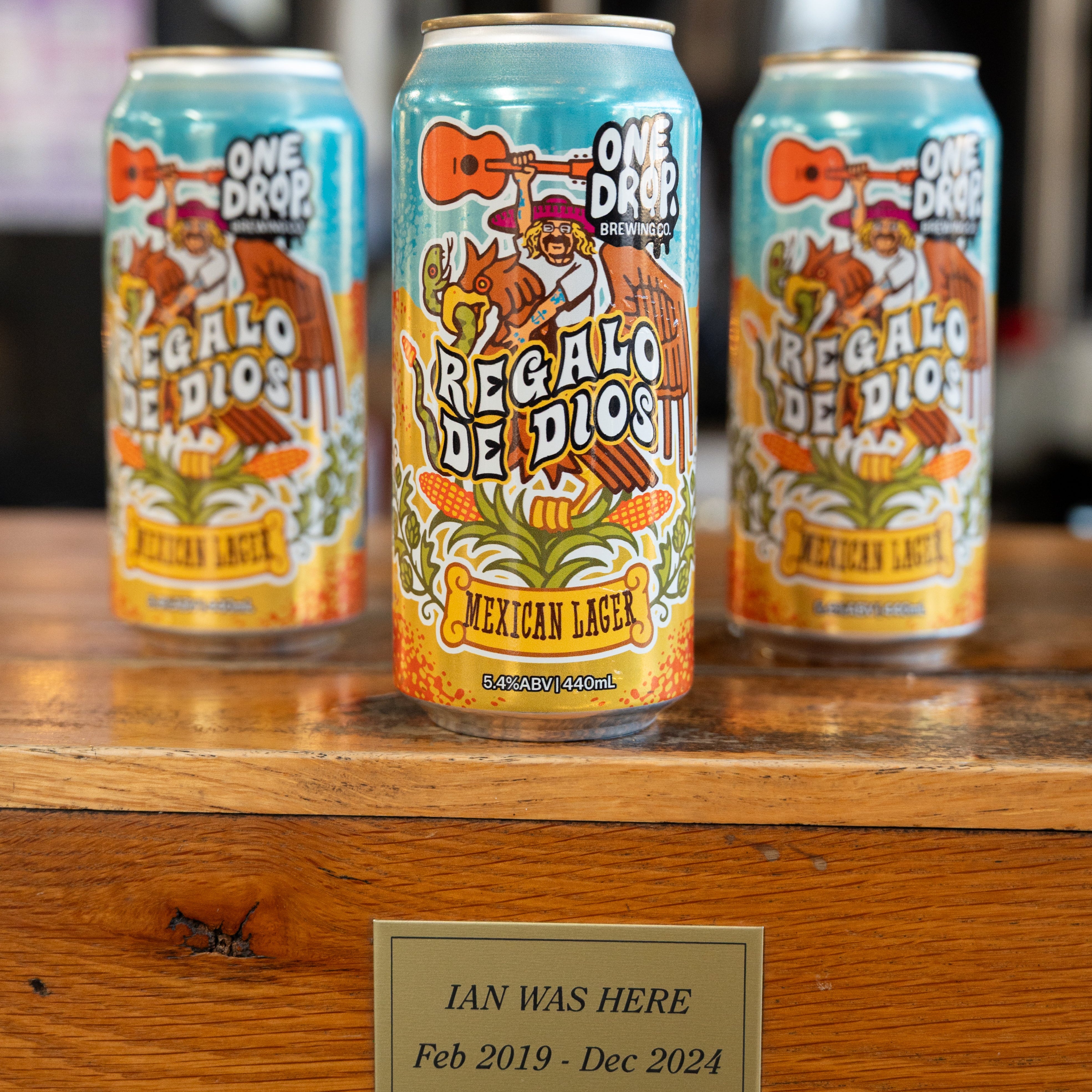 Three One Drop 'Regalo de Dios' Mexican Lager beer cans displayed on a wooden bar counter with a personalized name plaque in front.