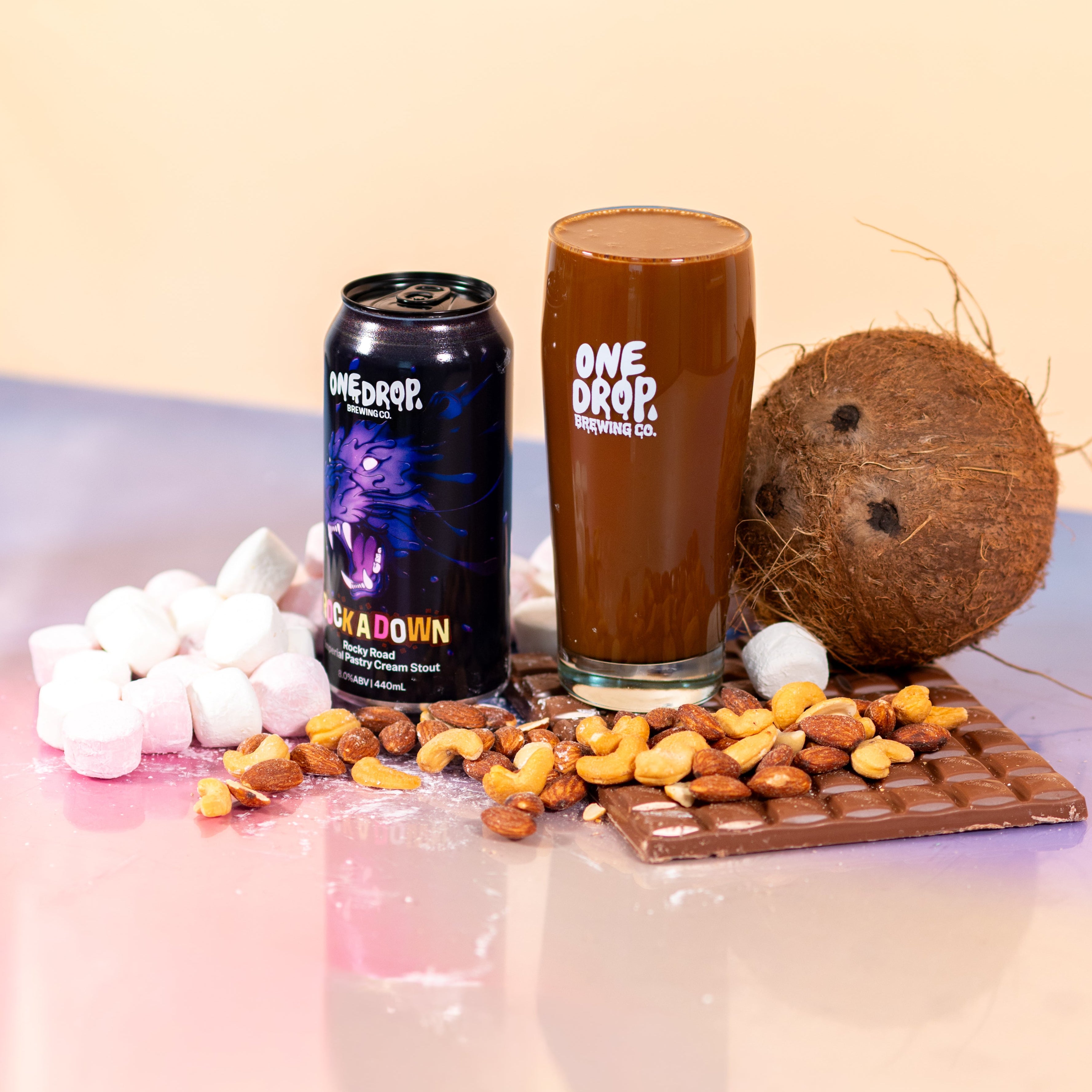 A glass of chocolate milk and a can of One Drop Chocolate 'Blackout' flavor, styled with a whole coconut, marshmallows, nuts, and a chocolate bar on a pastel pink and beige background.