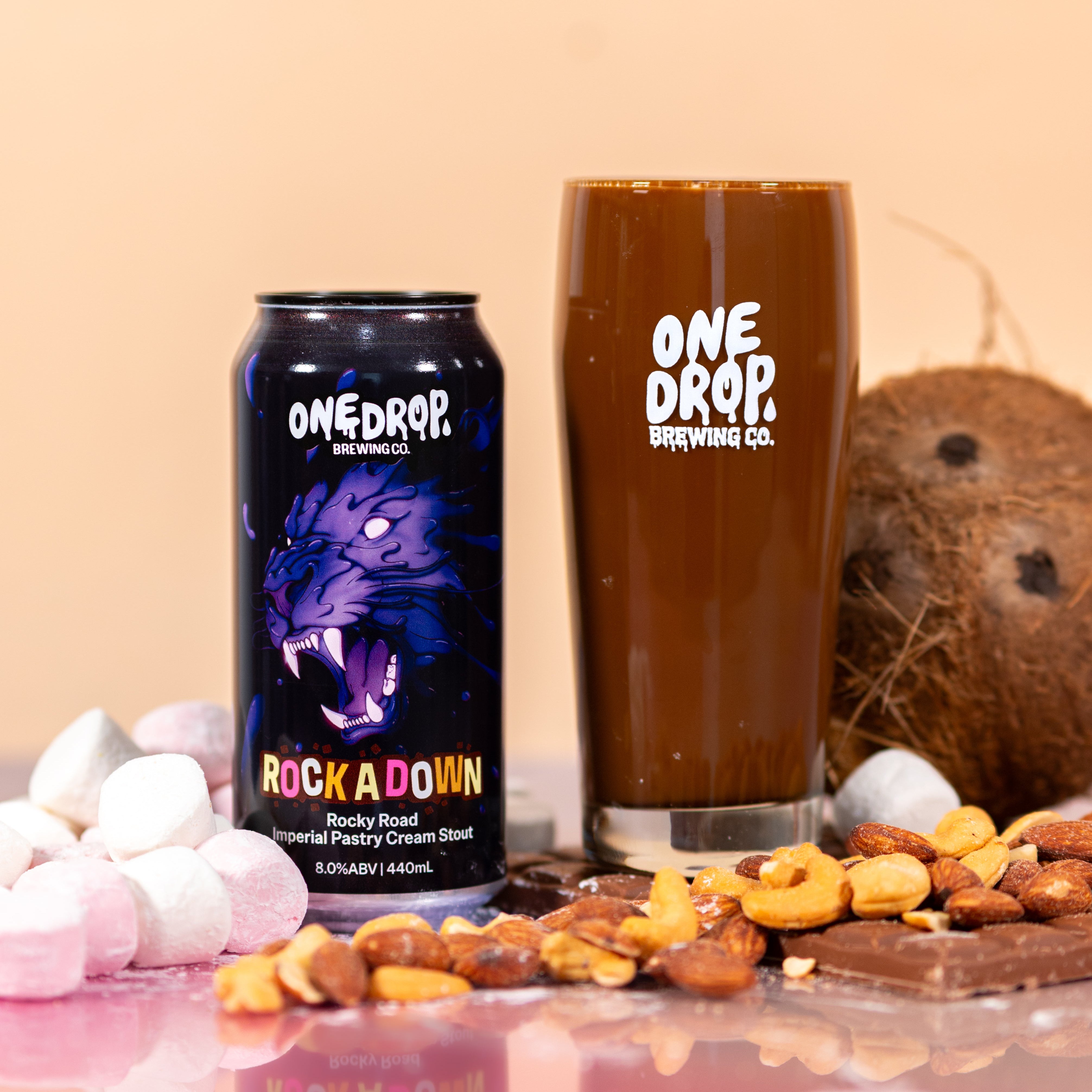 A can and a tall glass of One Drop Brewing Co. Rock A Down chocolate pastry cream stout, styled with chocolate, nuts, marshmallows, and a coconut on a pink reflective surface against a beige background.