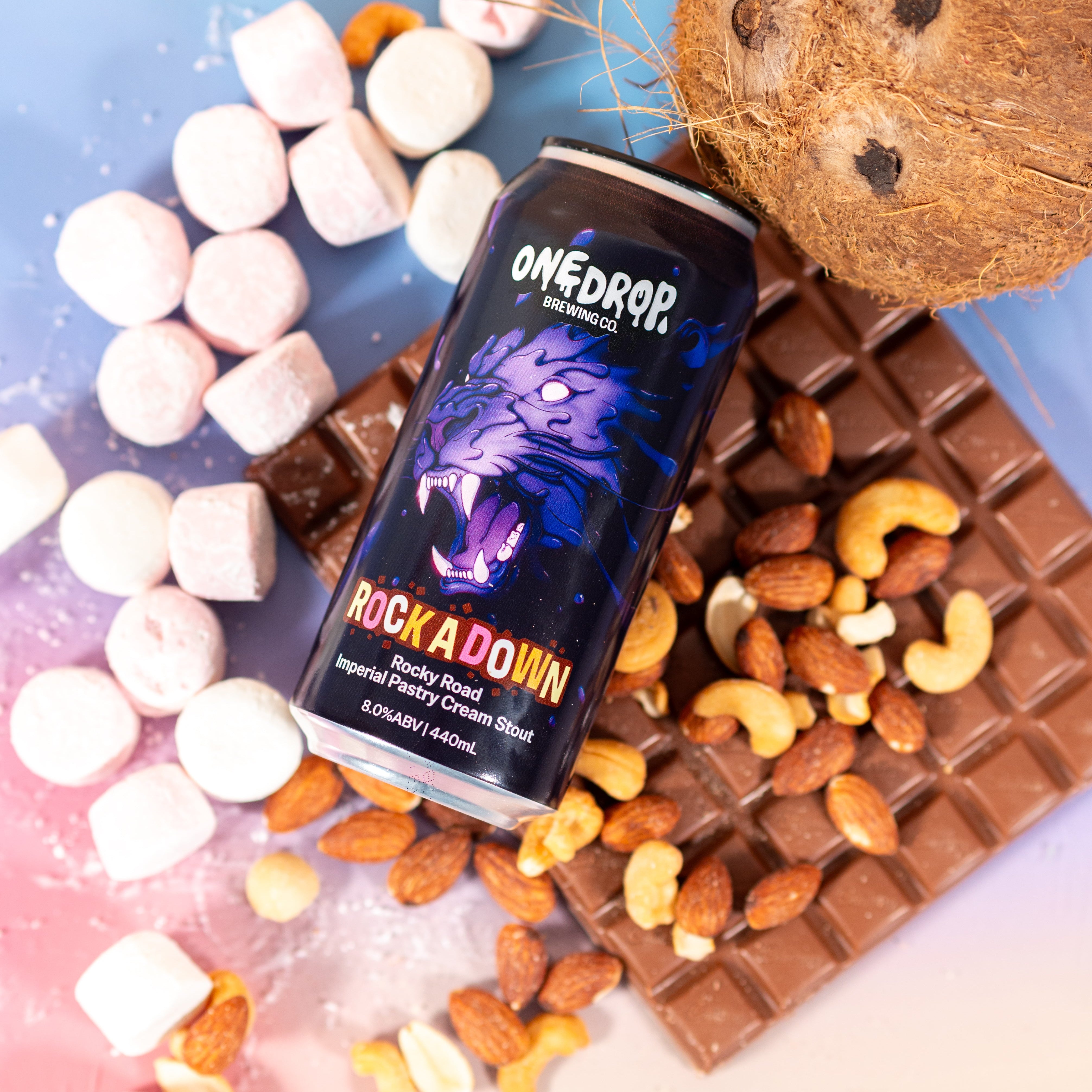 A can of OneDrop Rock A Down coconut-flavored malt beverage is styled in a flat-lay with a whole coconut, chocolate bar, almonds, cashews, and marshmallows on a pastel blue and pink gradient background.