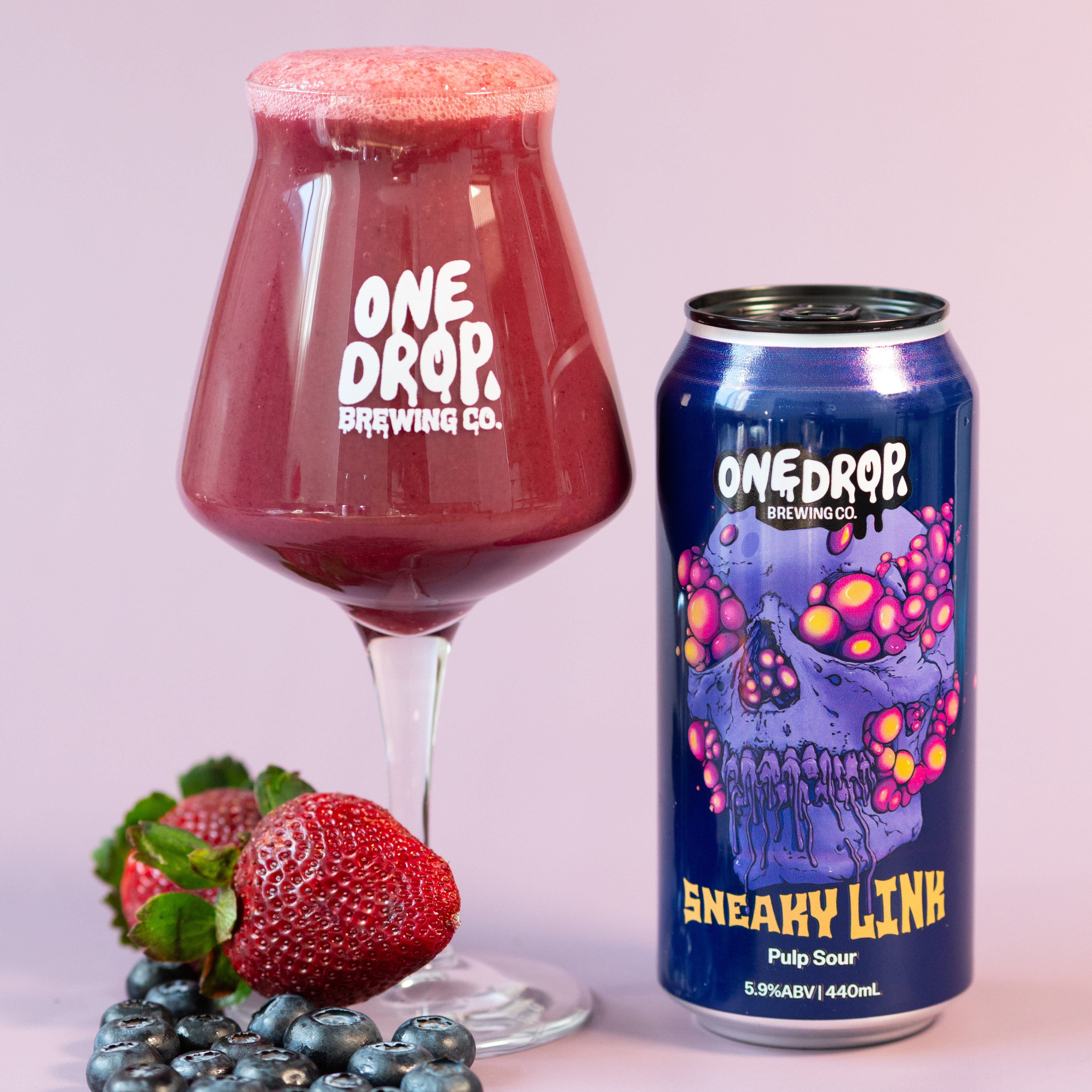 A glass of deep red beverage with a foamy head next to a blue can labeled One Drop Brewing Co. 'Sneaky Lick' Pulp Sour, styled with strawberries and blueberries on a soft pink background.