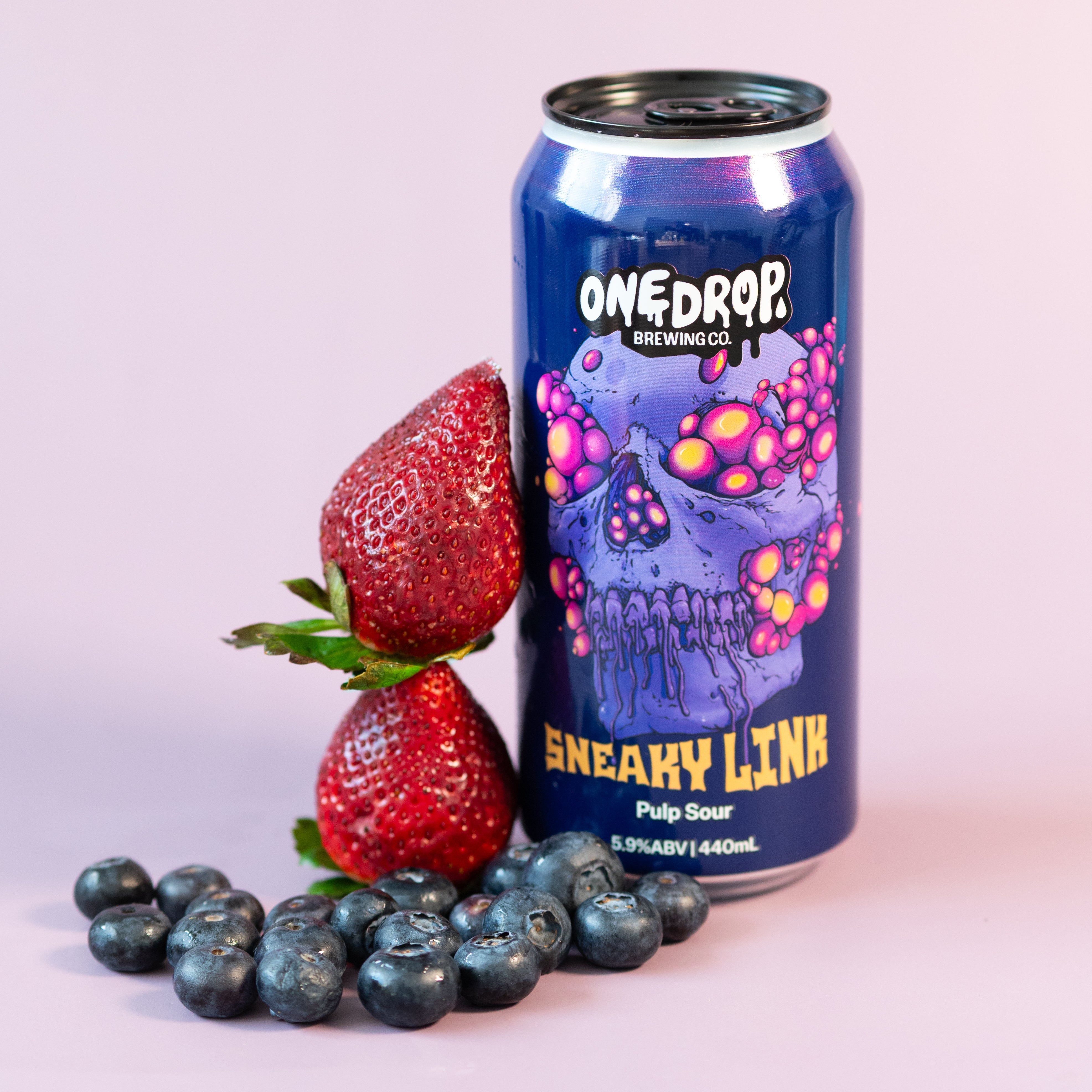 A can of OneDrop 'Sneaky Link' pulp sour beverage stands on a light pink background, styled with fresh strawberries and blueberries in the foreground.