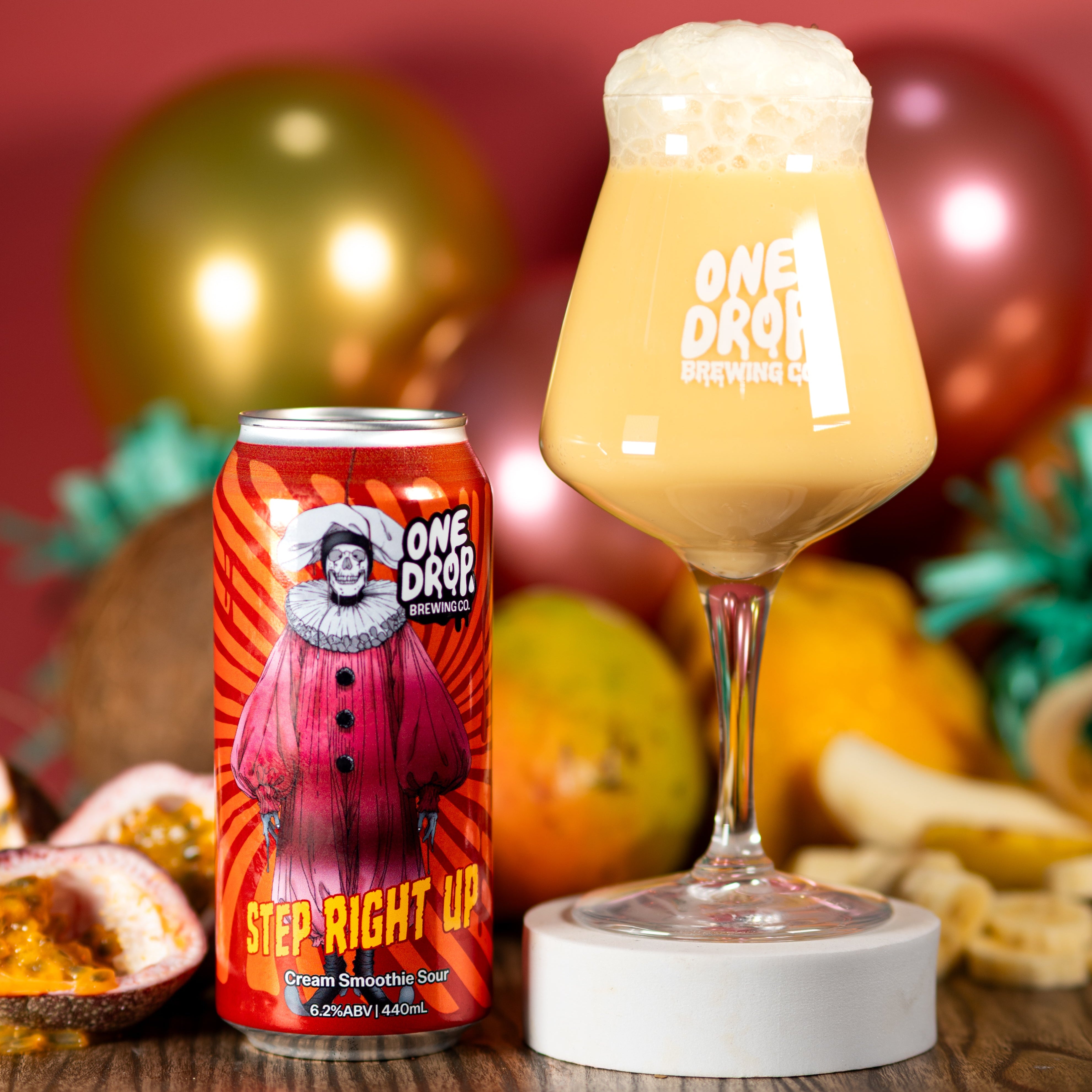 A can of One Drop Brewing 'Step Right Up' malt beverage next to a stemmed glass filled with the pale golden beer and topped with foam, set on a wooden surface with fruit and metallic balloons in the background.