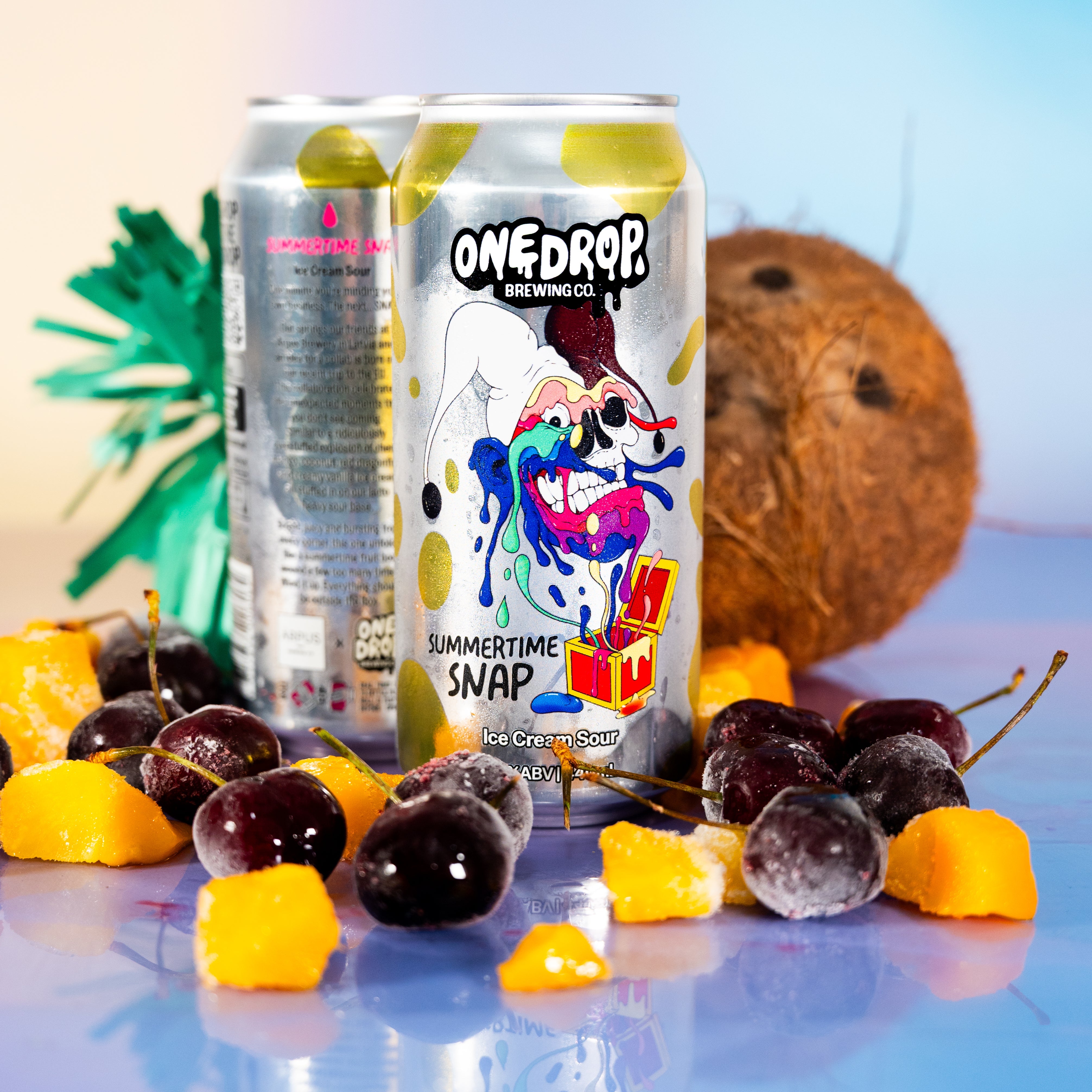 Two OneDrop Brewing Co. beverage cans standing upright on a glossy blue surface, surrounded by fruit slices and whole coconuts against a soft pastel gradient background.