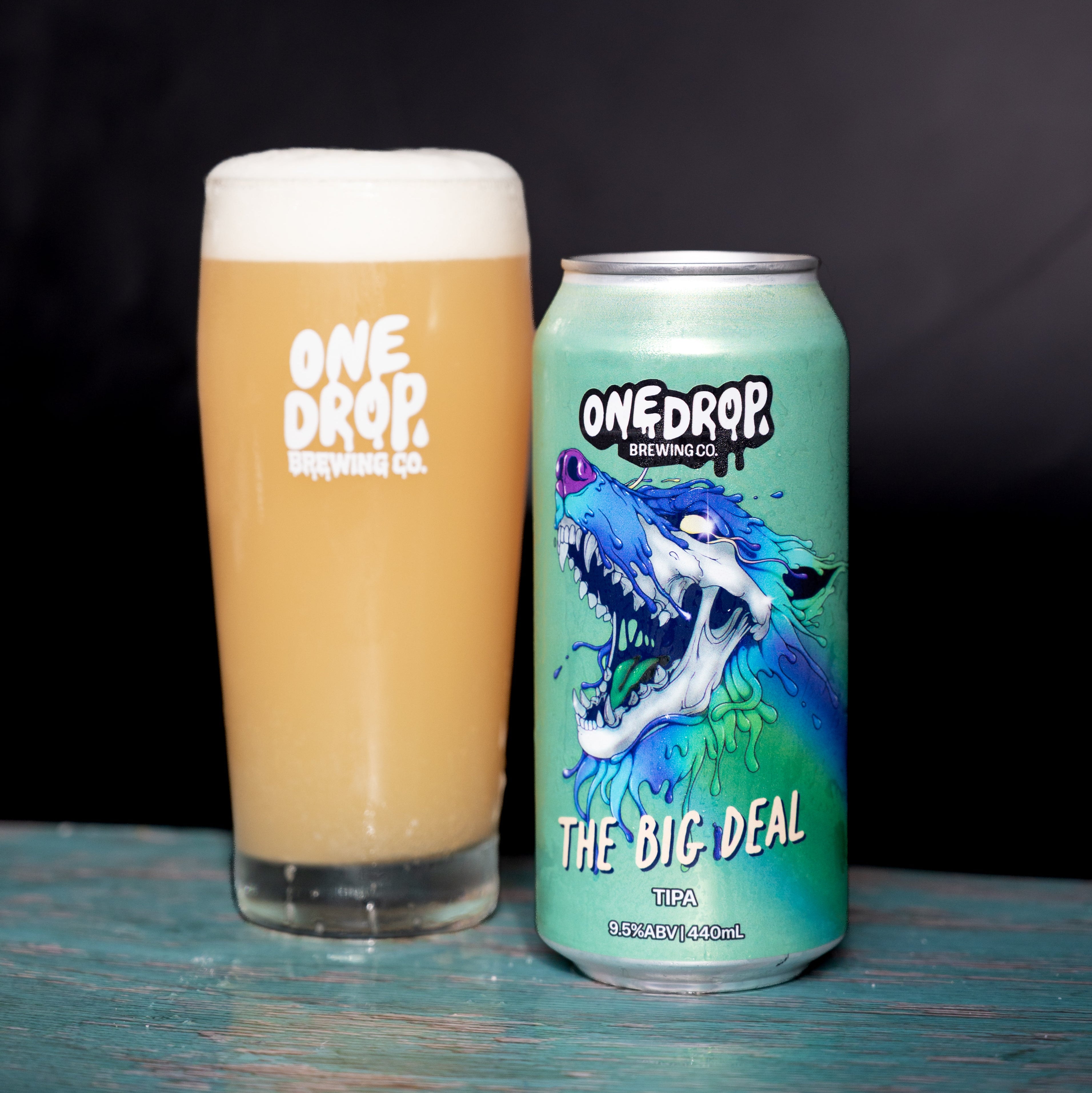 A glass of frothy golden beer and a can of One Drop Brewing Co. 'The Big Deal' IPA next to each other on a rustic blue wooden surface against a dark background.
