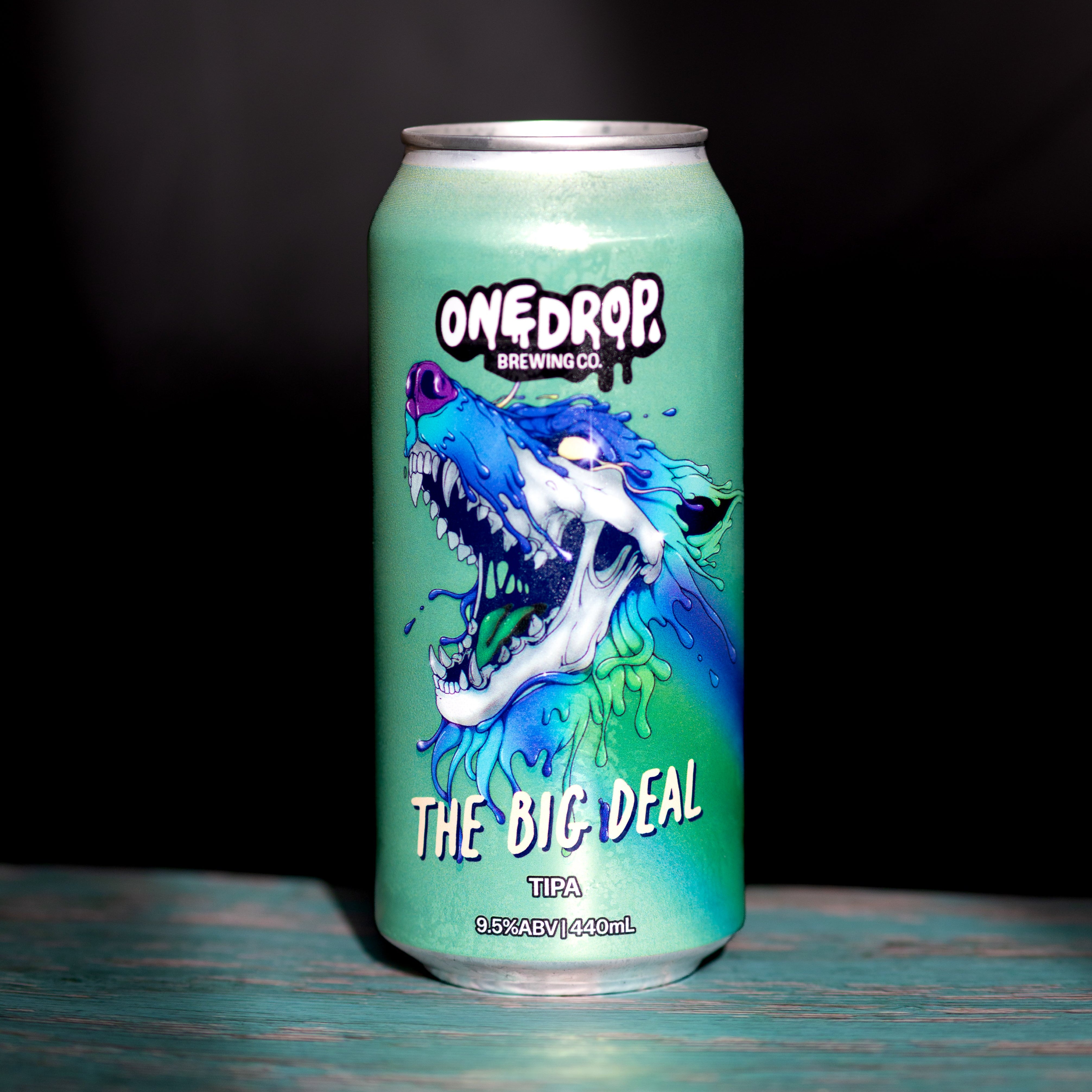 A 330ml aluminum can of OneDrop Brewing Co 'The Big Deal' IPA, featuring a vibrant green and blue wolf graphic, set on a rustic teal wooden surface against a dark background.