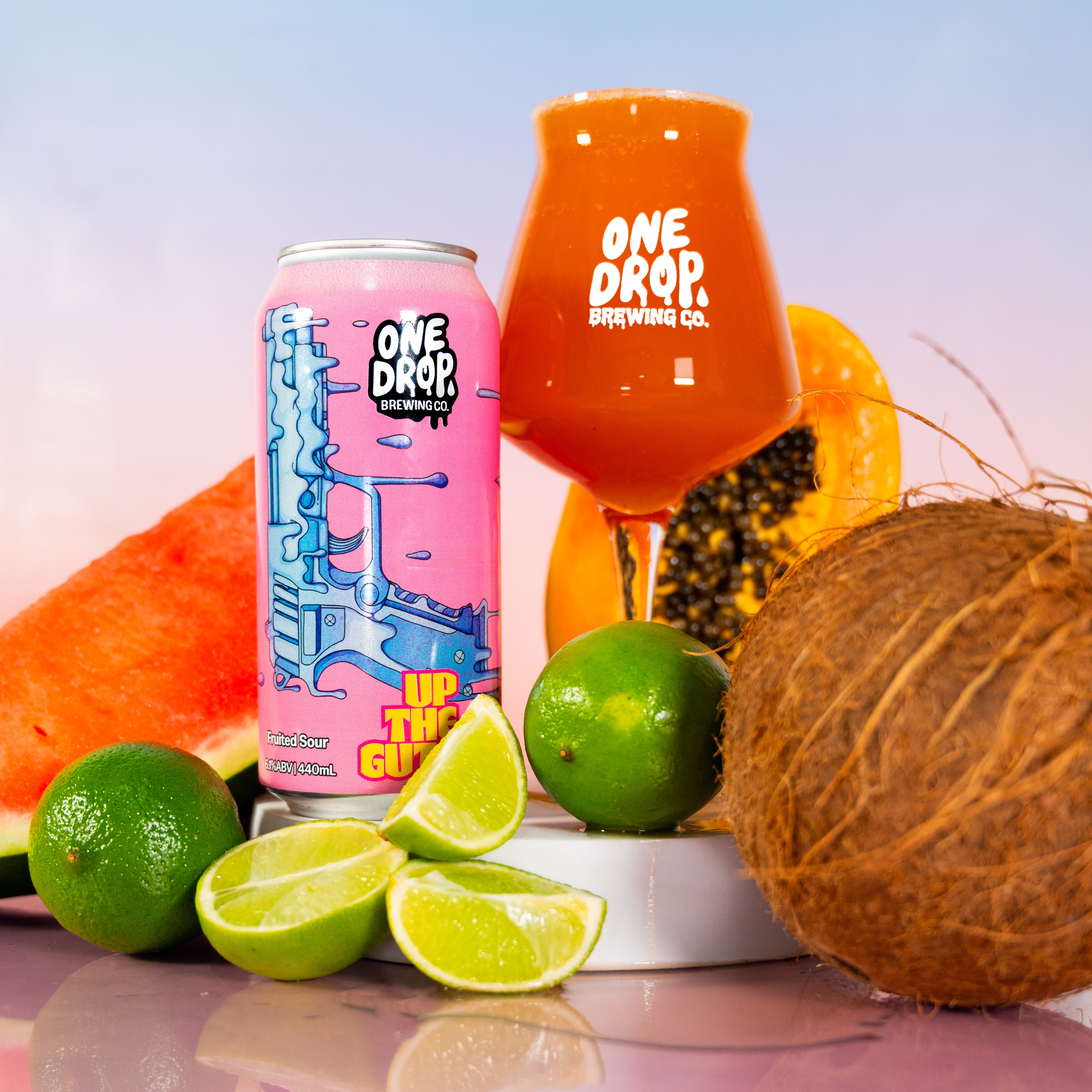 A beverage product display featuring a pink can and an orange-tinted glass both printed with the One Drop Brewing Co. logo, styled with limes, a papaya slice, a coconut, and a watermelon slice on a pink reflective surface against a pastel sky background.