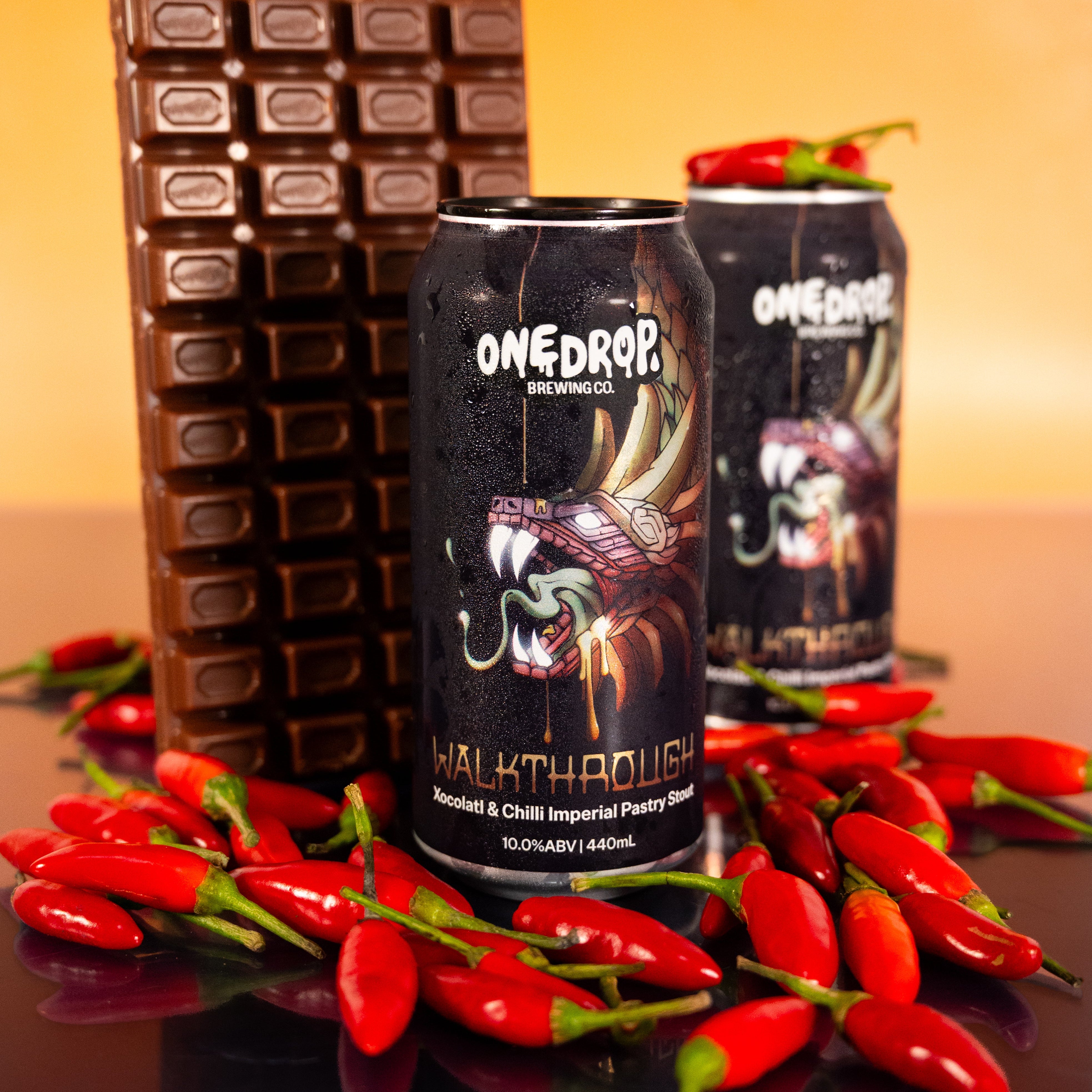 Two OneDrop Brews 'Walkthrouse' chili beer cans displayed on a reflective black surface, surrounded by fresh red chili peppers and a large chocolate bar, against a warm orange gradient background.