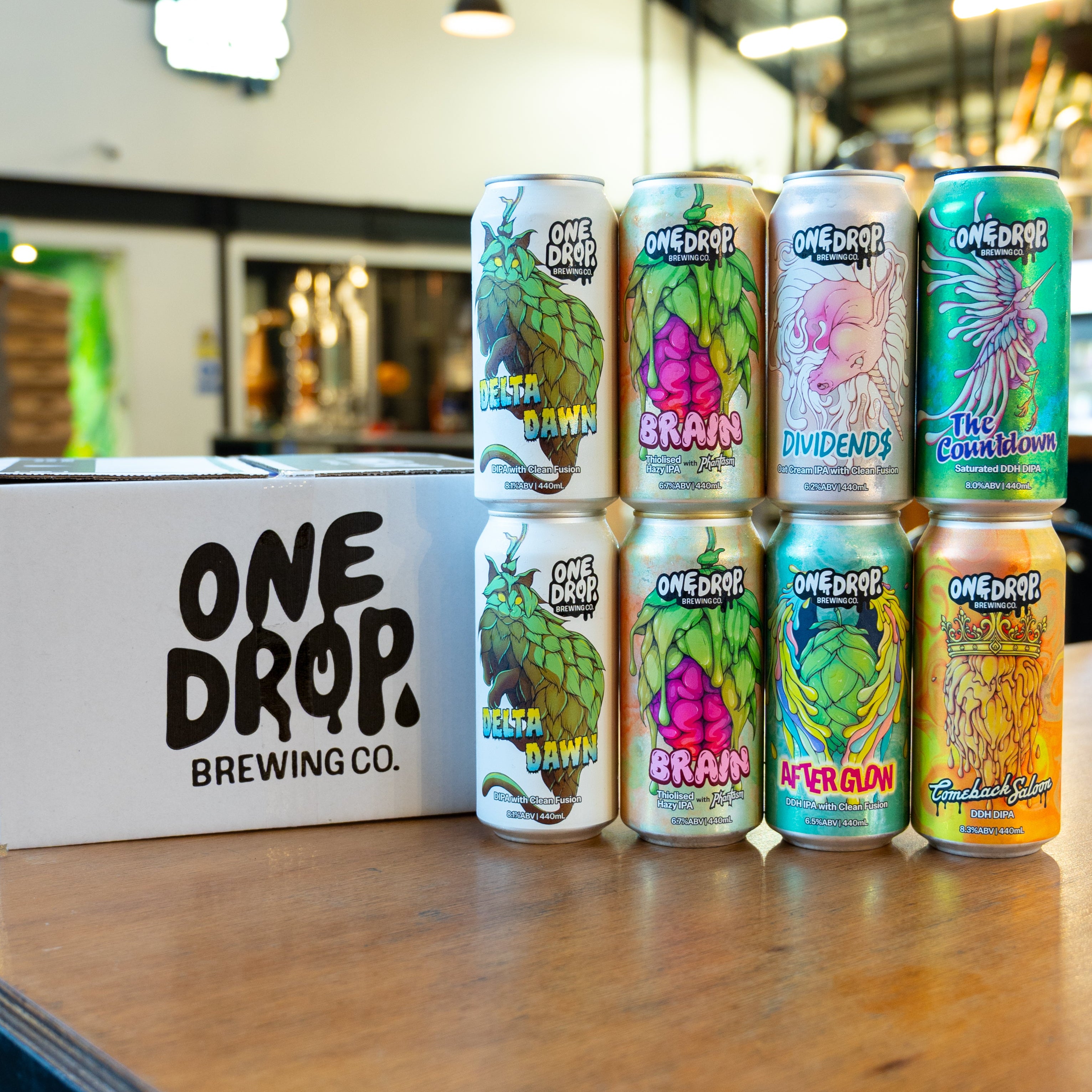 Assorted cans of One Drop Brewing Co. beer on a wooden table with a branded box.